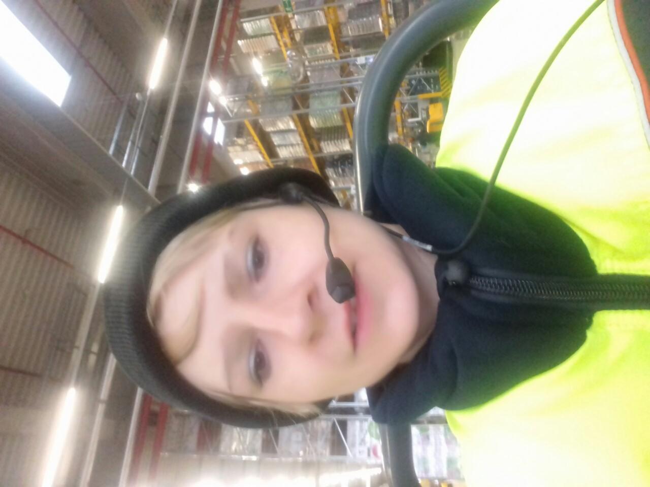 Pracownica w kamizelce odblaskowej i słuchawkach na tle magazynu. Widok z dołu, oświetlenie jarzeniowe.