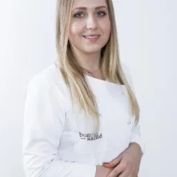 Portret uśmiechniętej blondynki w białym uniformie medycznym z logo 'bonne santé' na kieszeni, dłonie splecione przed sobą, na jasnym tle.