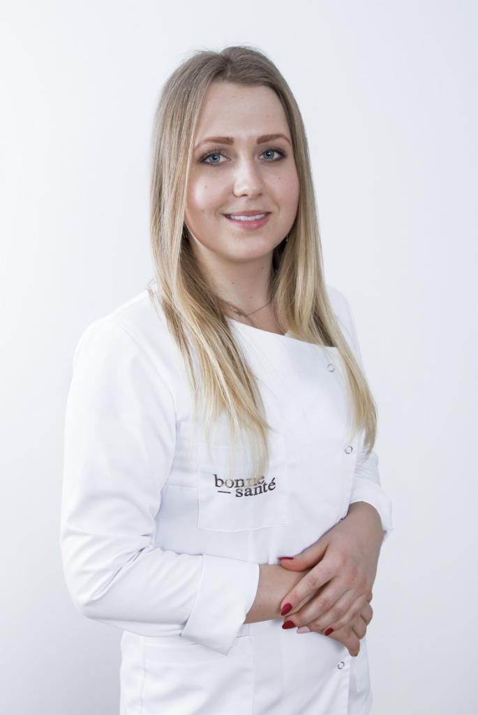 Portret uśmiechniętej blondynki w białym uniformie medycznym z logo 'bonne santé' na kieszeni, dłonie splecione przed sobą, na jasnym tle.