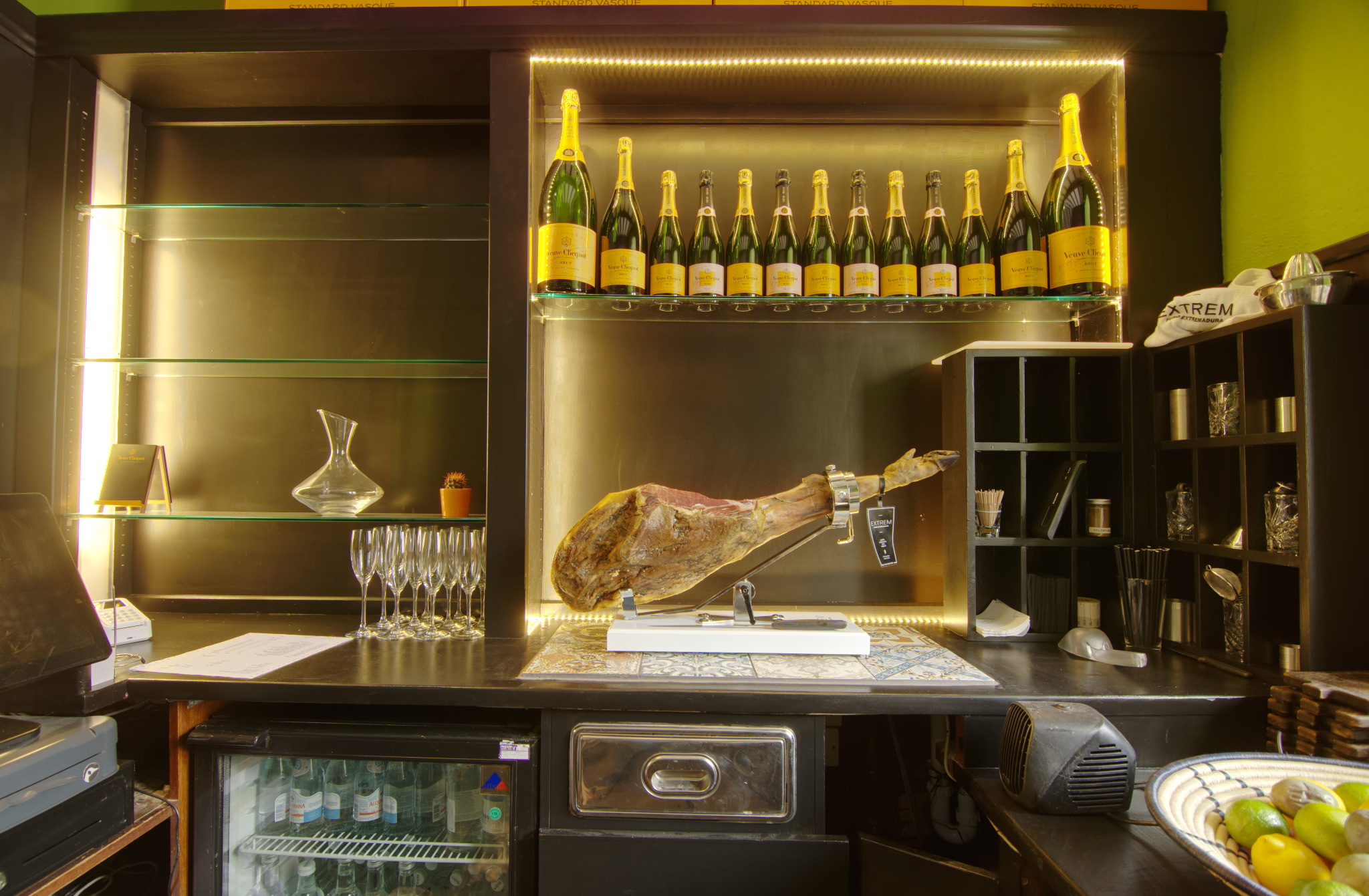 Bar z szynką Jamon na stojaku, butelkami szampana Veuve Clicquot na górnej półce, karafką i kieliszkami do szampana po lewej, oraz lodówką z napojami pod blatem.