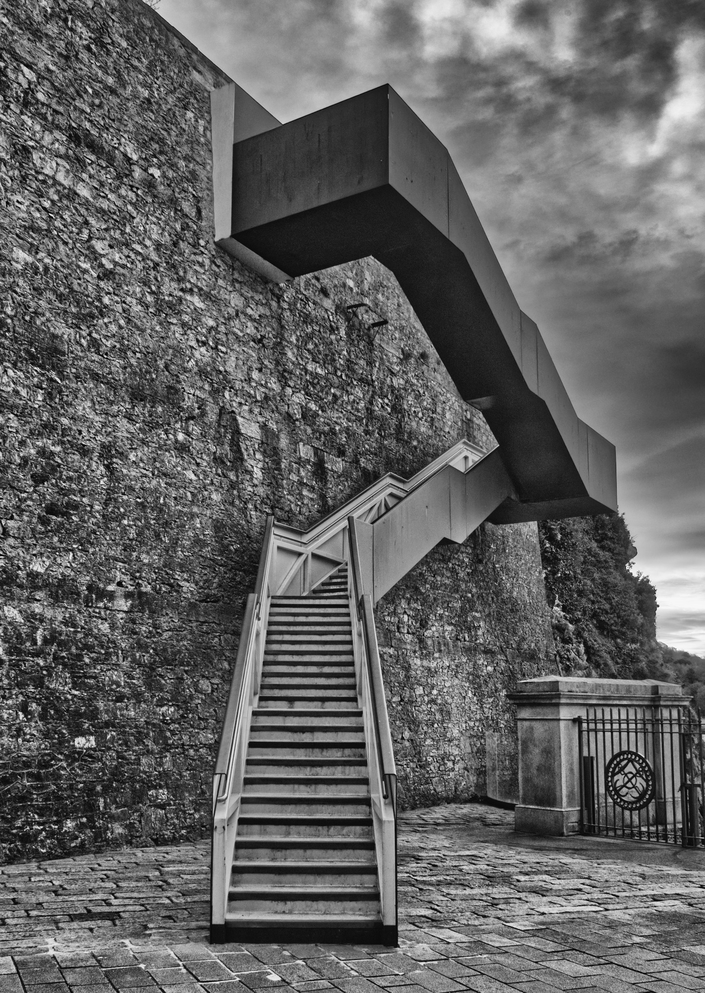Czarno-białe zdjęcie zewnętrznych schodów prowadzących wzdłuż kamiennej ściany z nowoczesną, betonową konstrukcją nad nimi, zakończonych bramą z ozdobnym motywem.