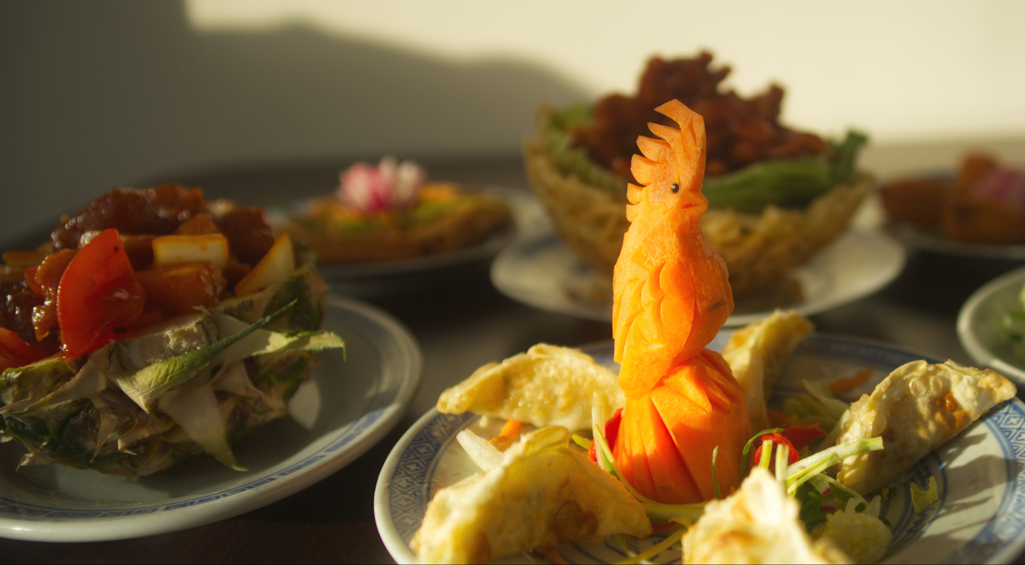 Dekoracja stołu z rzeźbą w marchewce przedstawiającą koguta, pierożkami i potrawą z ananasem w tle, na talerzach z niebieskim wzorem.