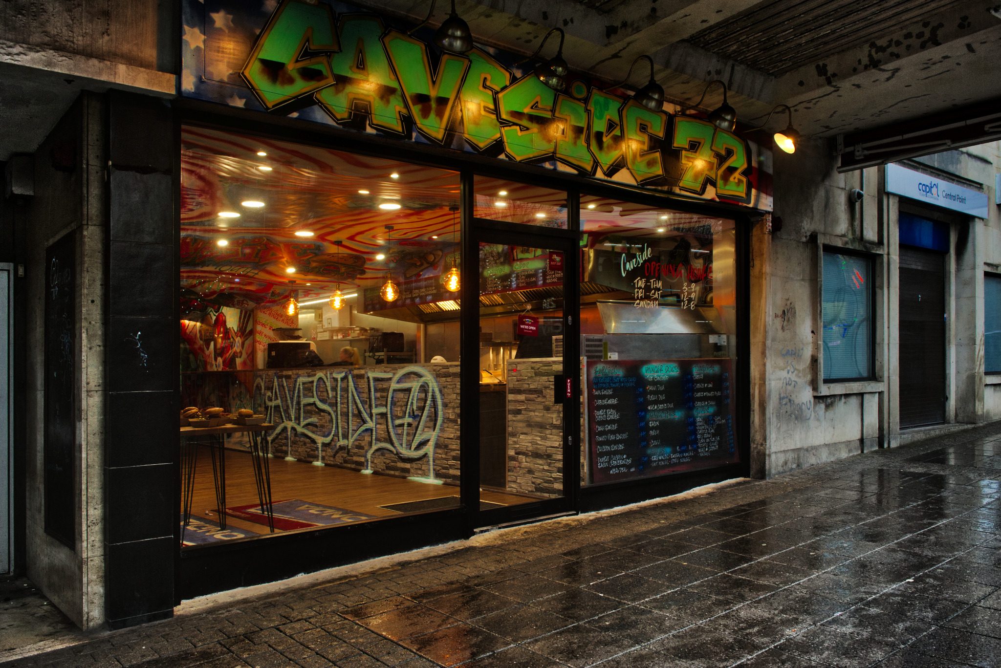 Fasada lokalu gastronomicznego z graffiti w stylu ulicznym, widoczna przez szybę menu wypisane kredą i wnętrze z dekoracyjnym malowaniem sufitu.