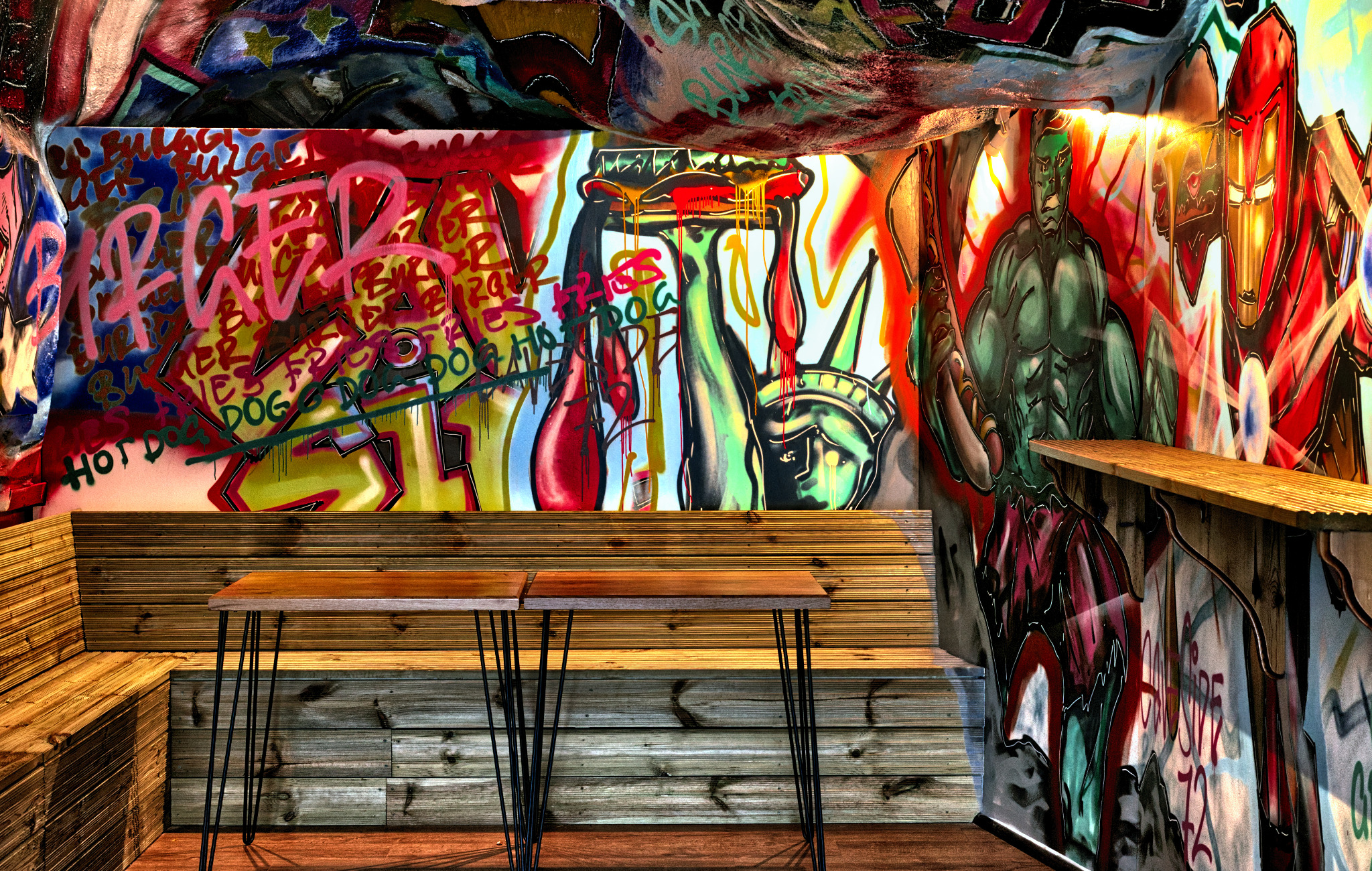 Wnętrze lokalu z drewnianą ławą i stolikiem na metalowych nogach, ściany pokryte kolorowymi graffiti przedstawiającymi Statuę Wolności, Hulka i Iron Mana oraz napisy. Sufit również pokryty malunkami.