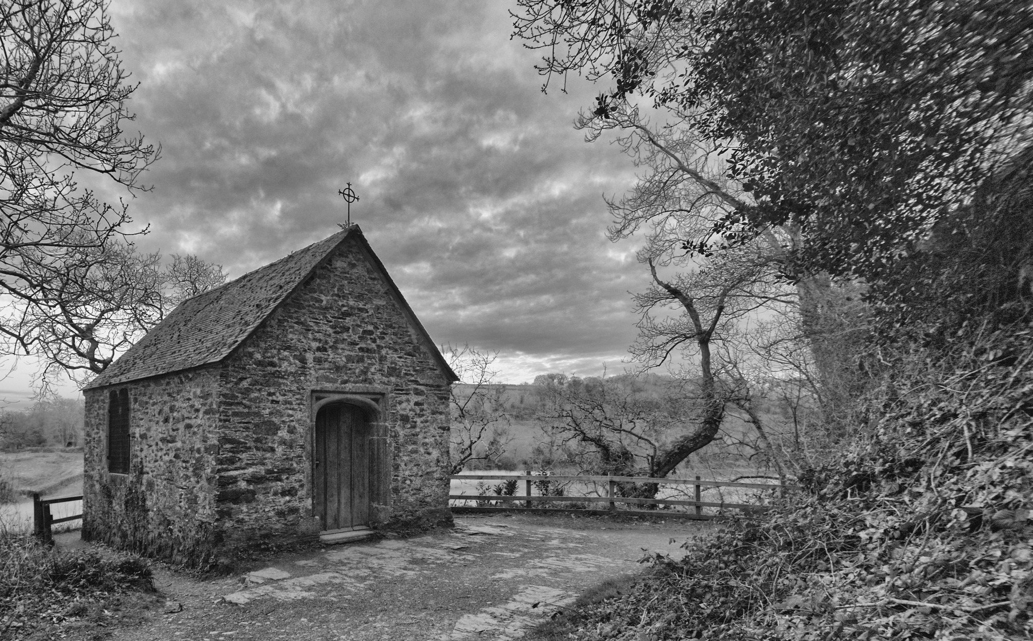 Czarno-białe zdjęcie kamiennej kaplicy z krzyżem na dachu, otoczonej drzewami i krzewami, pod pochmurnym niebem.