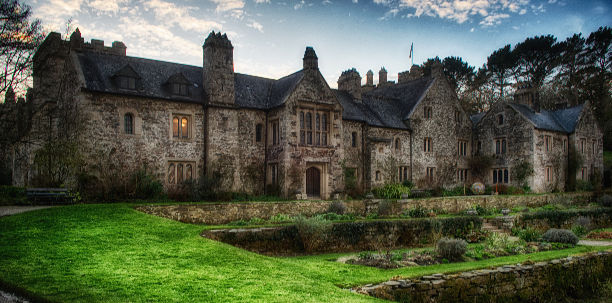 Rozległy, kamienny dwór z wieloma oknami i stromymi dachami, otoczony zielonym trawnikiem i kamiennymi murkami. Okno na pierwszym planie świeci ciepłym, pomarańczowym światłem.