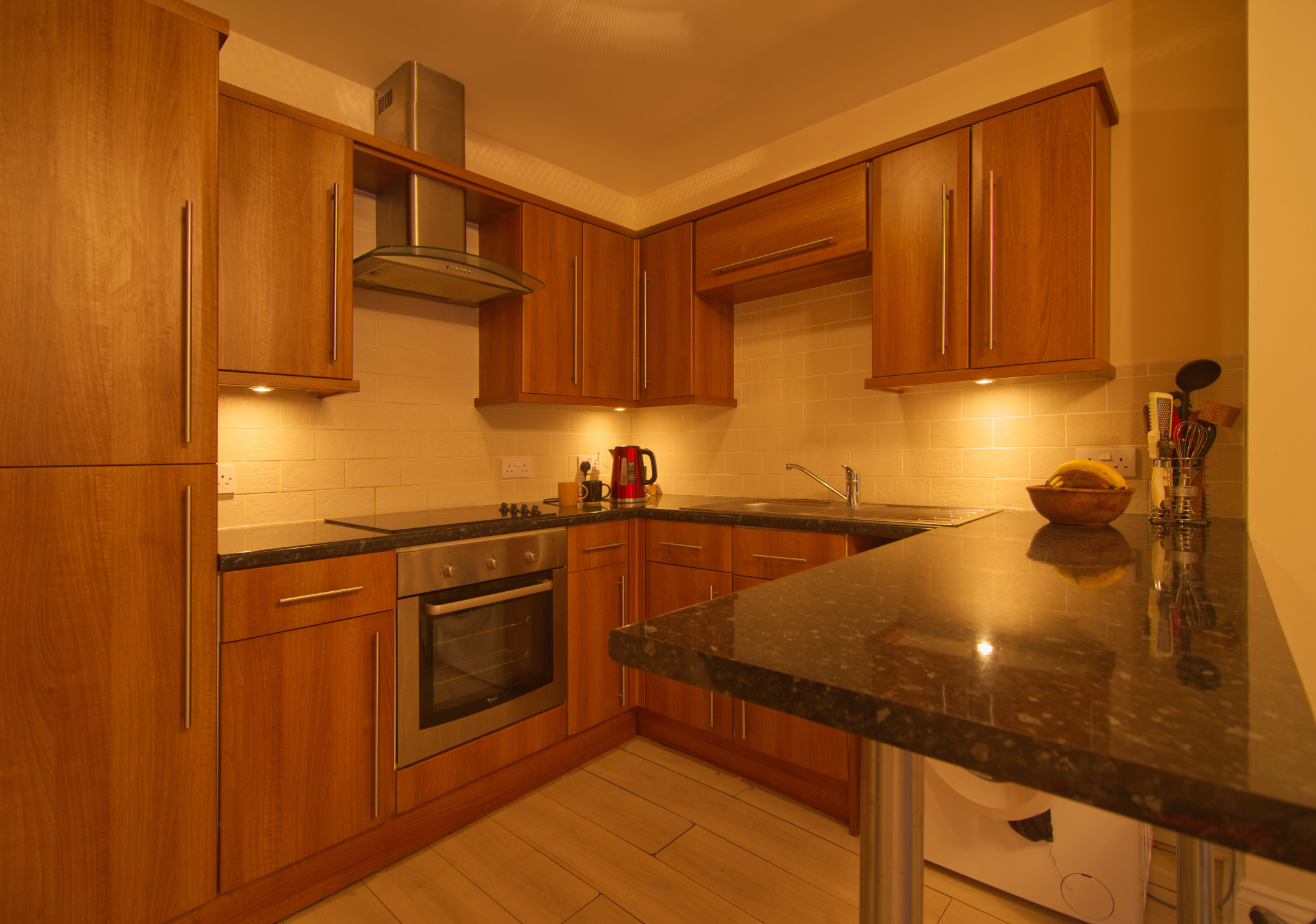 Kuchnia z drewnianymi szafkami, czarnym blatem i sprzętami AGD, oświetlona ciepłym światłem. Na blacie misa z owocami i czerwony czajnik.