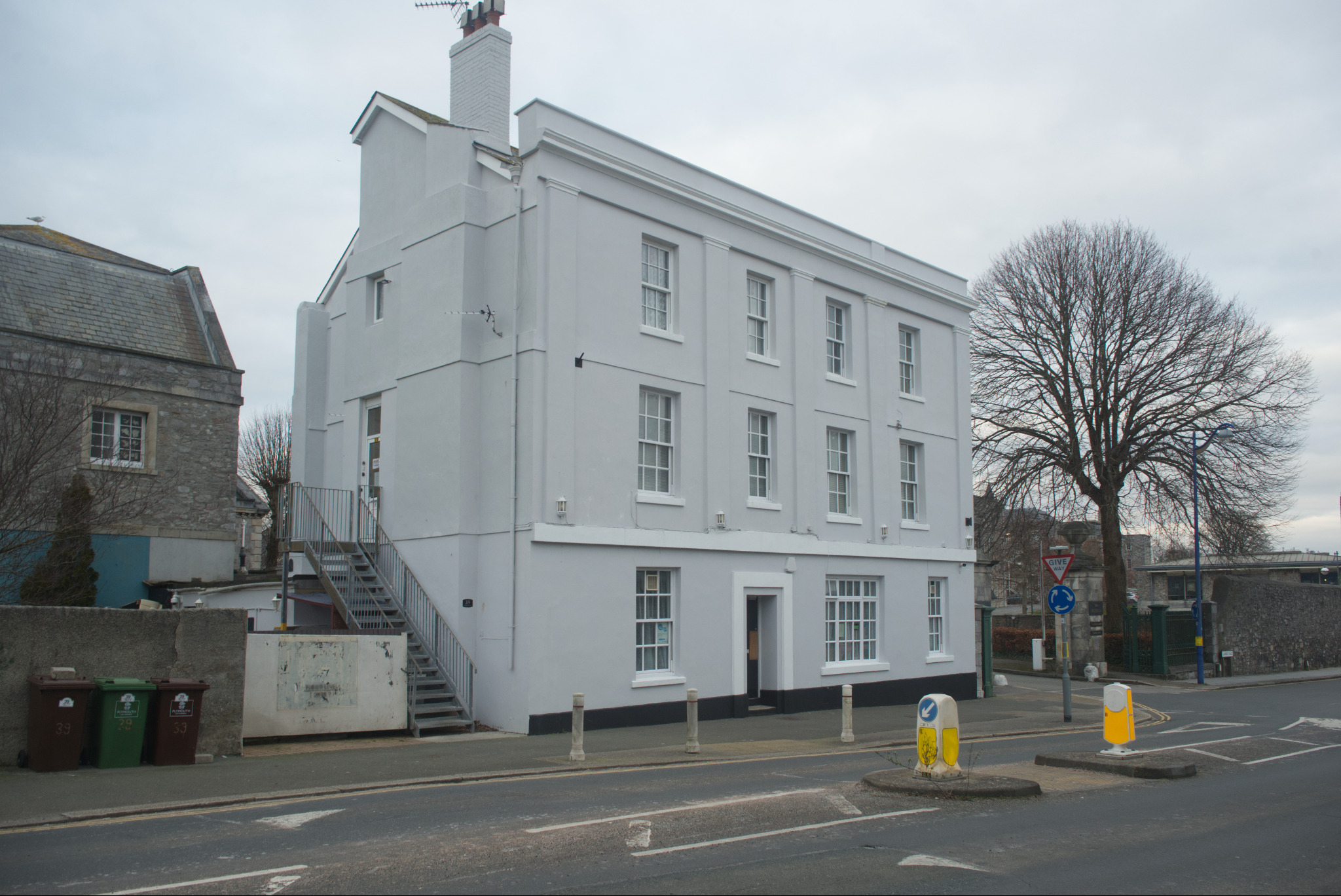 Odnowiony, trzypiętrowy budynek pomalowany na szaro, z zewnętrznymi schodami prowadzącymi na pierwsze piętro i białymi obramowaniami okien