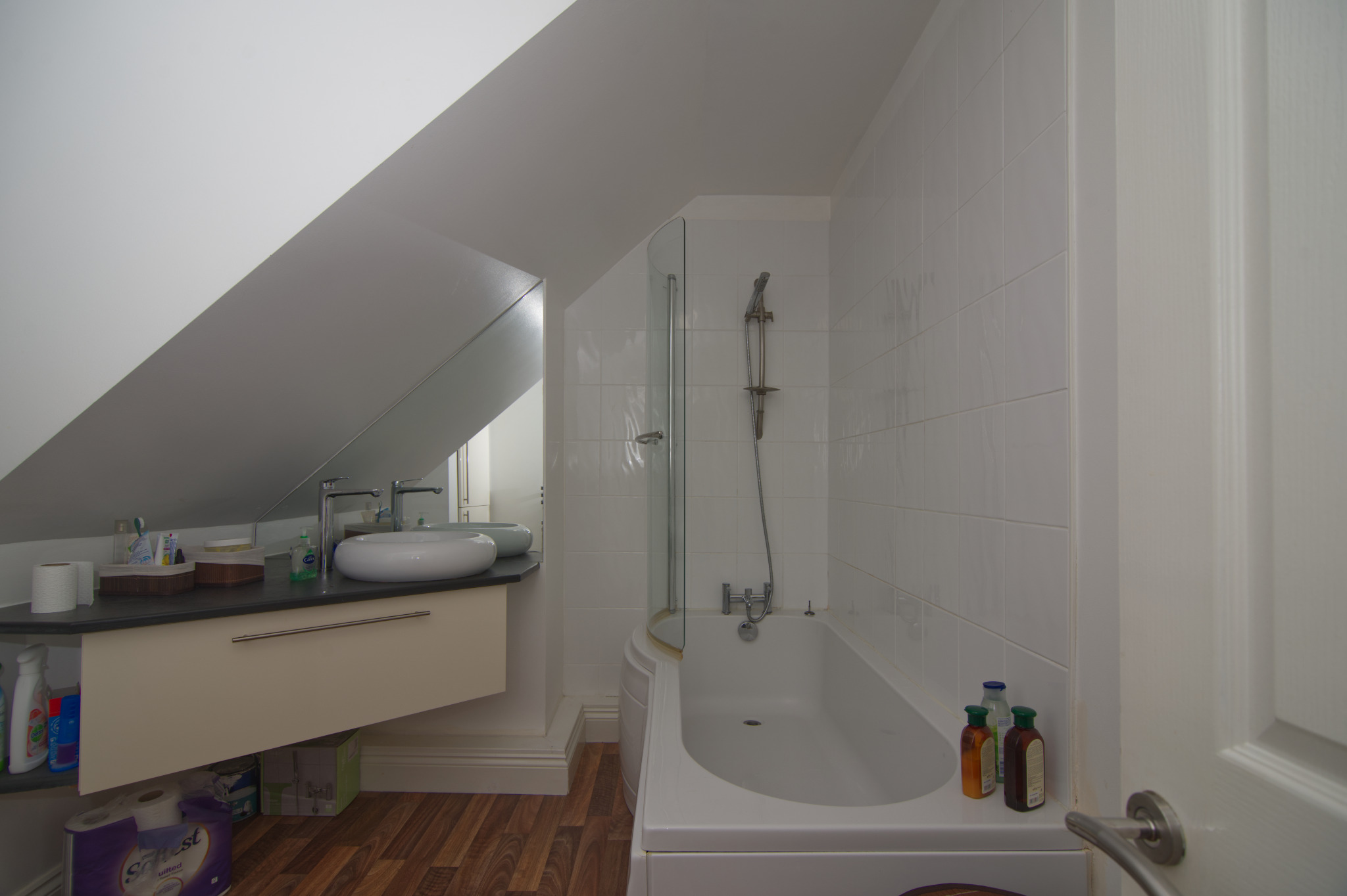 Jasna łazienka na poddaszu ze skośnym sufitem, z białą wanną z kabiną prysznicową, umywalką nablatową i drewnopodobną podłogą.