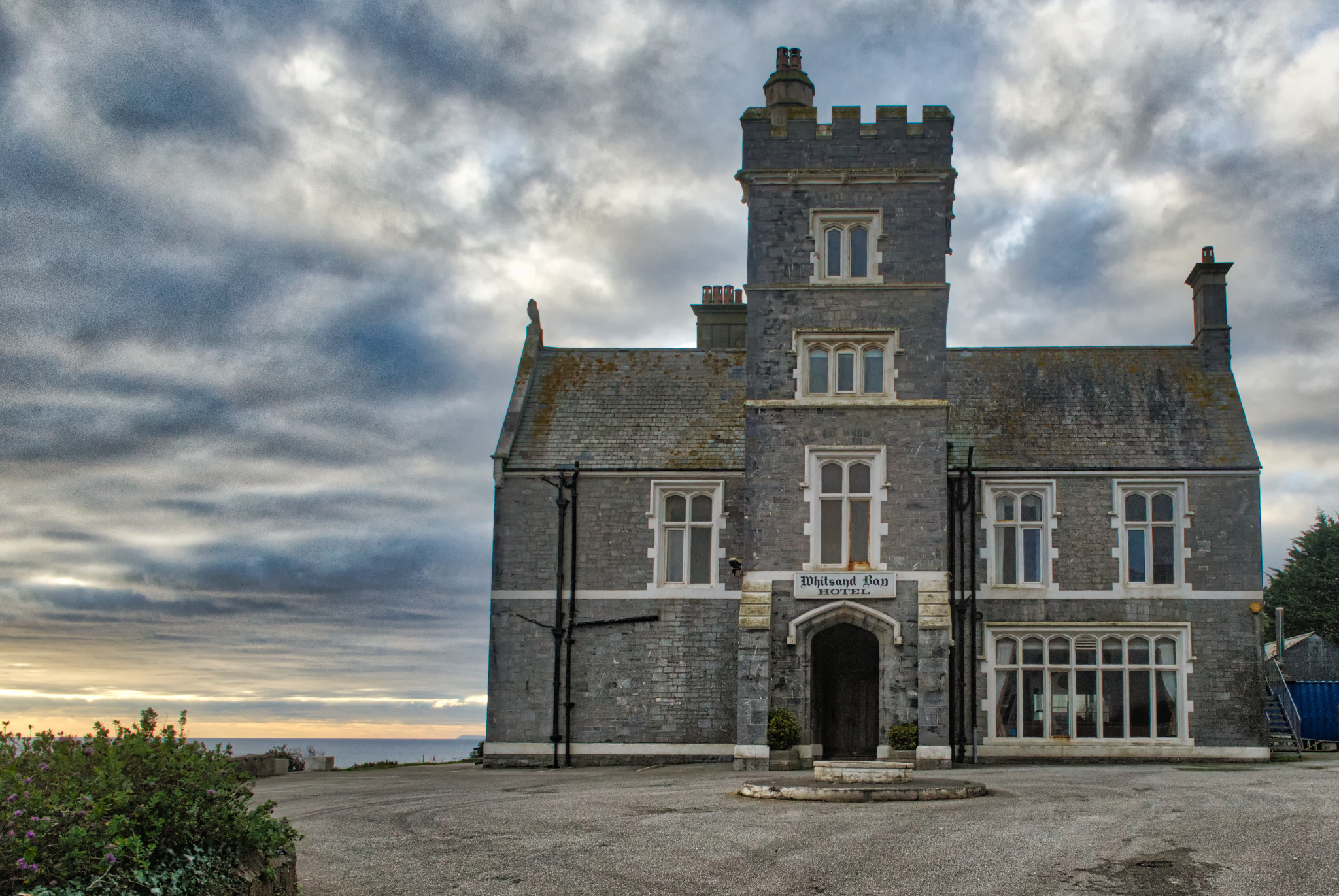 Elewacja kamiennego hotelu z wieżą i napisem 'Whitsand Bay Hotel' nad wejściem, na tle pochmurnego nieba i fragmentu morza.