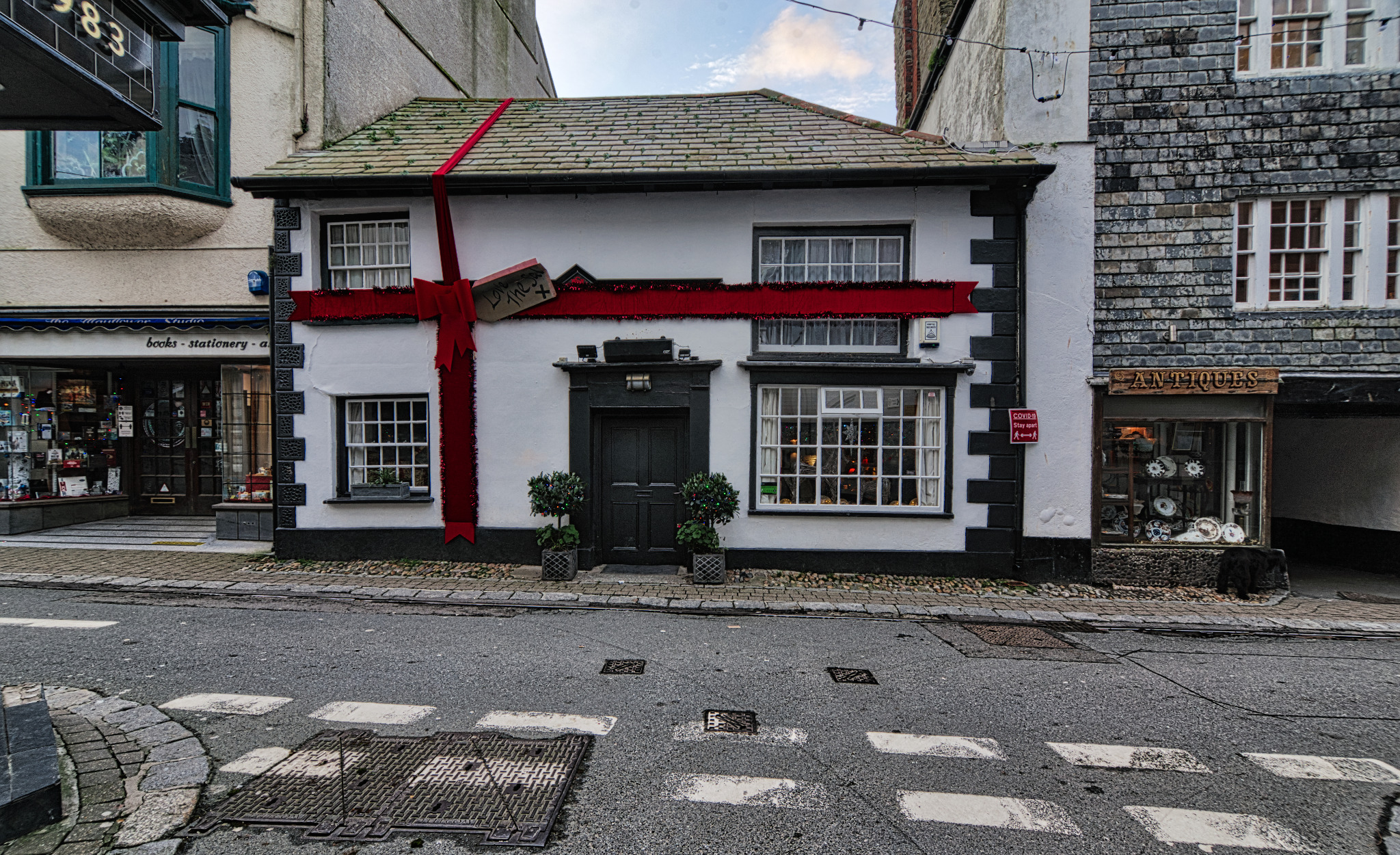 Niski budynek udekorowany jak prezent świąteczny z czerwoną wstążką, stojący między sklepem z książkami a antykami, widok z ulicy.