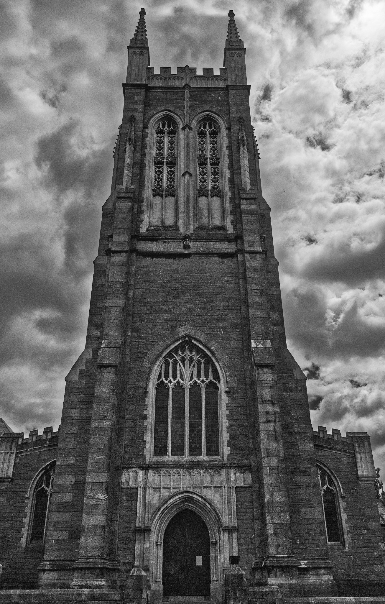Czarno-białe ujęcie wieży kościelnej z gotyckimi oknami i detalami architektonicznymi, sfotografowanej z dołu na tle burzowych chmur.