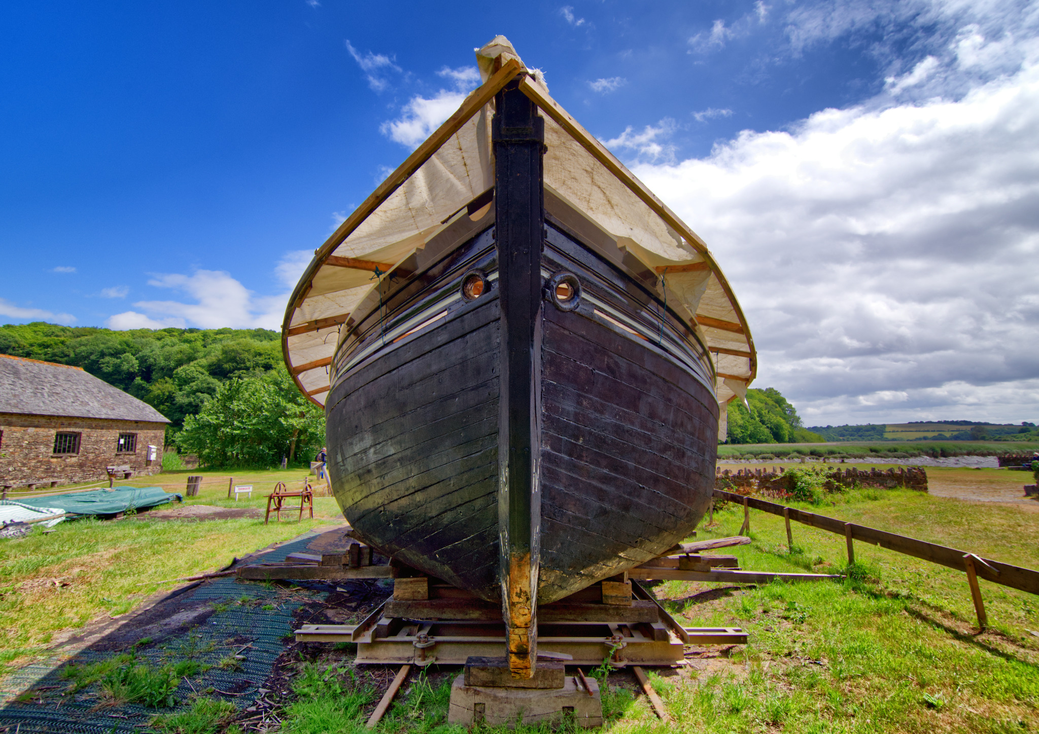 Frontowy widok klasycznej drewnianej łodzi na drewnianych podporach, z płóciennym zadaszeniem, na trawiastym terenie z budynkiem w tle pod błękitnym niebem z chmurami.