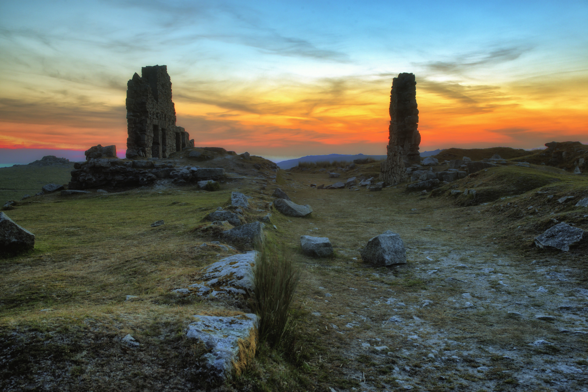 Ruiny kamiennej budowli z dwoma filarami na wzgórzu, widoczne na tle pomarańczowo-żółtego zachodu słońca z niebieskim niebem u góry.