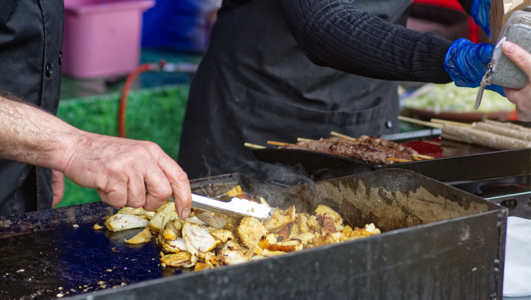 Przygotowywanie mięsa na grillu: kucharz w czarnym fartuchu obraca szczypcami kawałki kurczaka na rozgrzanej płycie, obok leżą szaszłyki na patyku, klient w niebieskich rękawiczkach płaci za...