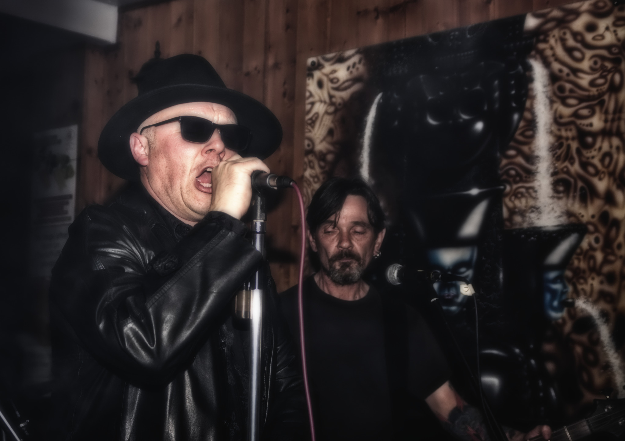 Wokalista w czarnym kapeluszu i skórzanej kurtce śpiewa do mikrofonu podczas koncertu, za nim gitarzysta i abstrakcyjne malowidło ścienne w odcieniach brązu i czerni.