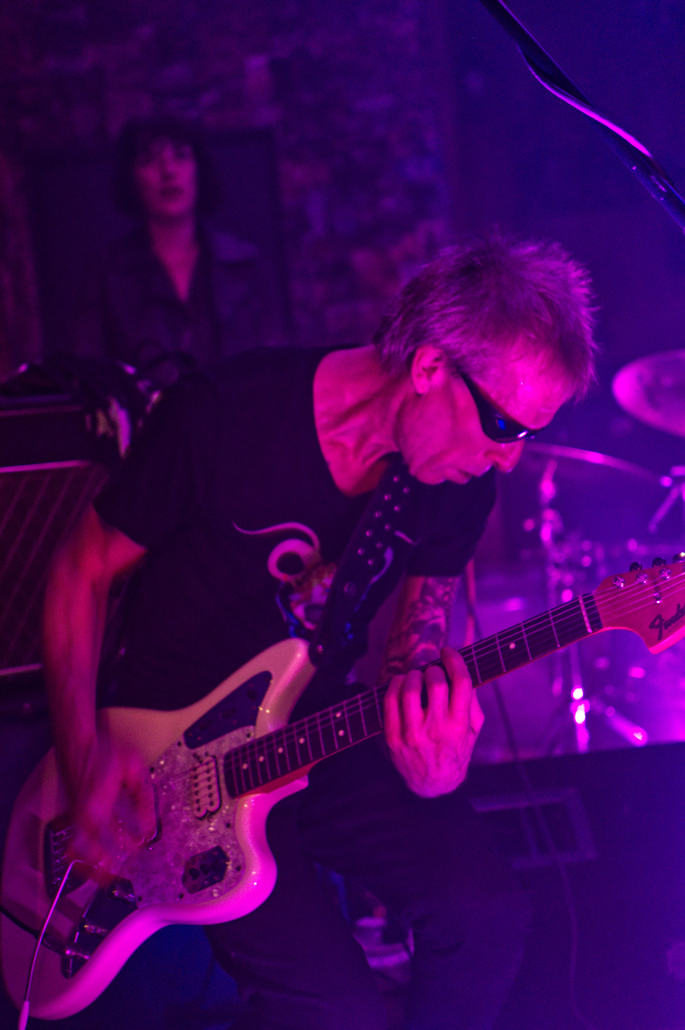 Muzyk grający na białej gitarze Fender Jaguar w fioletowym oświetleniu scenicznym, z widocznymi tatuażami na ramieniu i publicznością w tle.