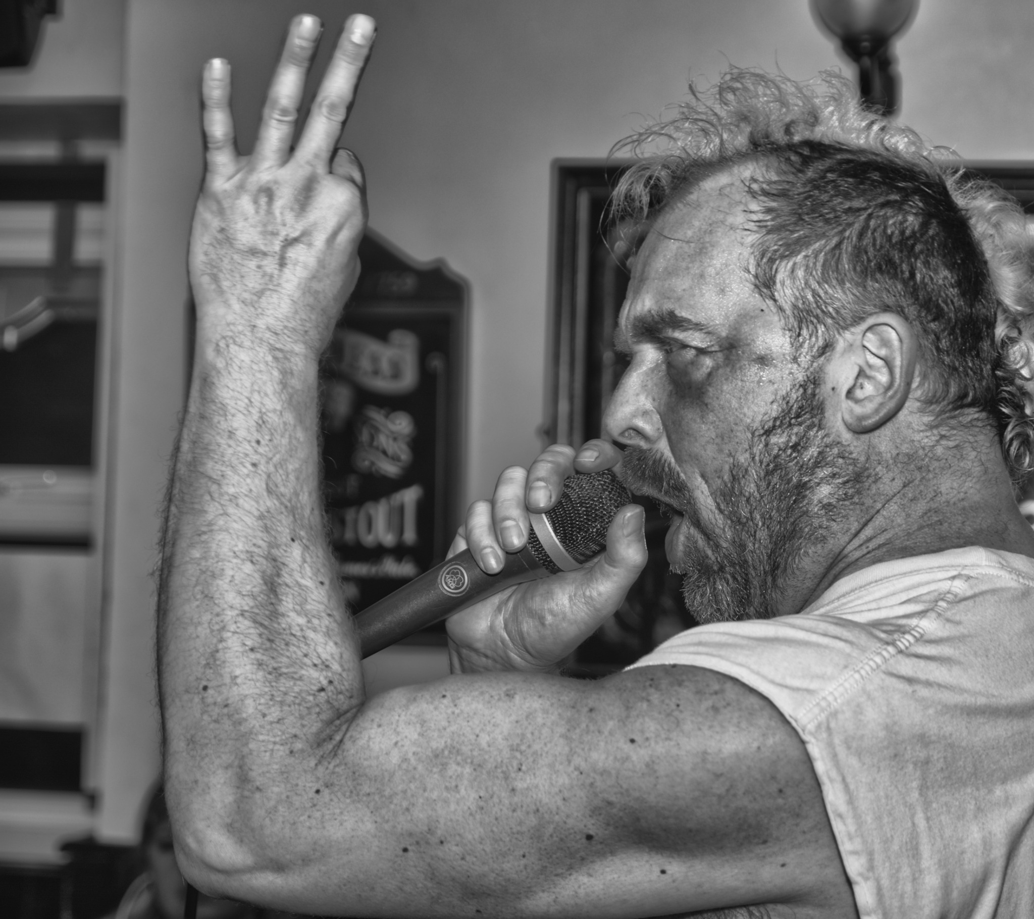 Czarno-białe zdjęcie profilu mężczyzny z brodą, śpiewającego do mikrofonu i unoszącego rękę z trzema palcami w górę, w tle widoczna tablica z napisem.