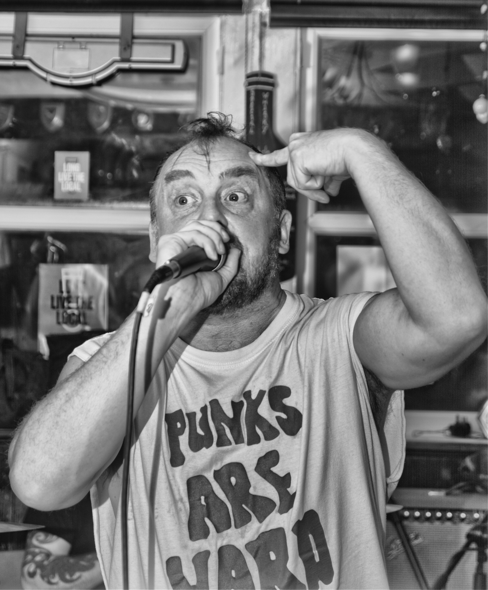 Czarno-białe zdjęcie mężczyzny z mikrofonem, wskazującego palcem na głowę, w tle okno z napisem 'Live the local', na koszulce napis 'Punks are hard'.