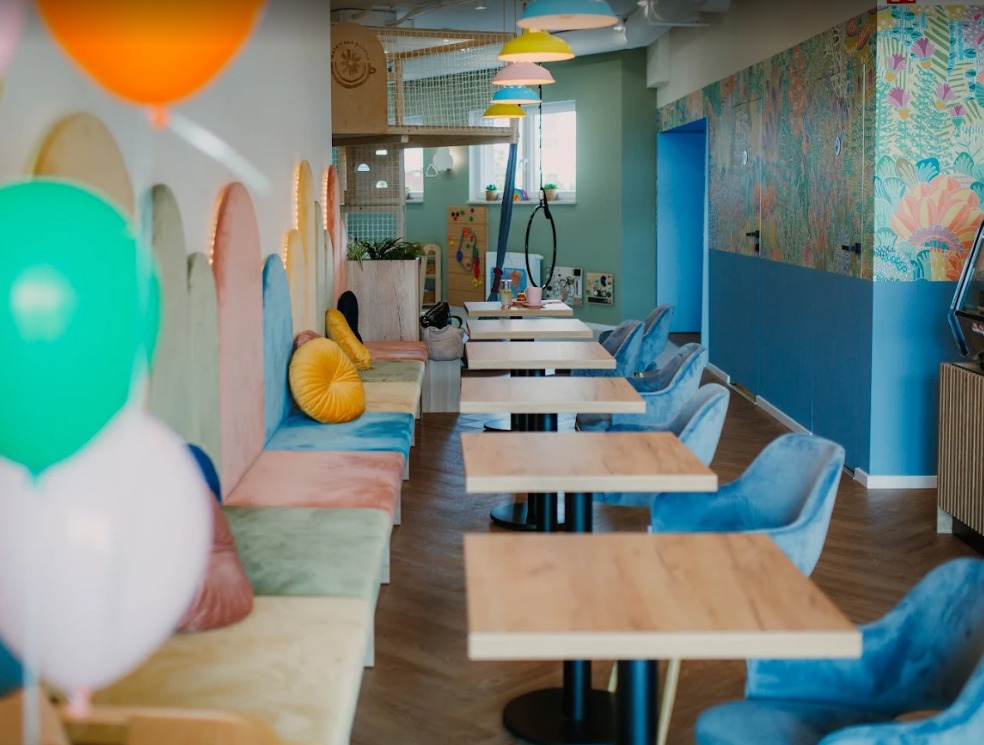 Pastelowe wnętrze sali zabaw dla dzieci z drewnianymi stolikami, pluszowymi krzesłami i kolorowymi balonami, zaprojektowane przez architekta wnętrz.