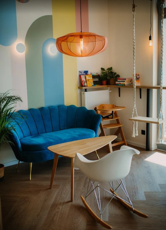 Stylowy pokój z turkusową sofą, drewnianym stolikiem i białym fotelem na biegunach. Na ścianie kolorowe pasy i designerska lampa. W tle huśtawka i półka z książkami.