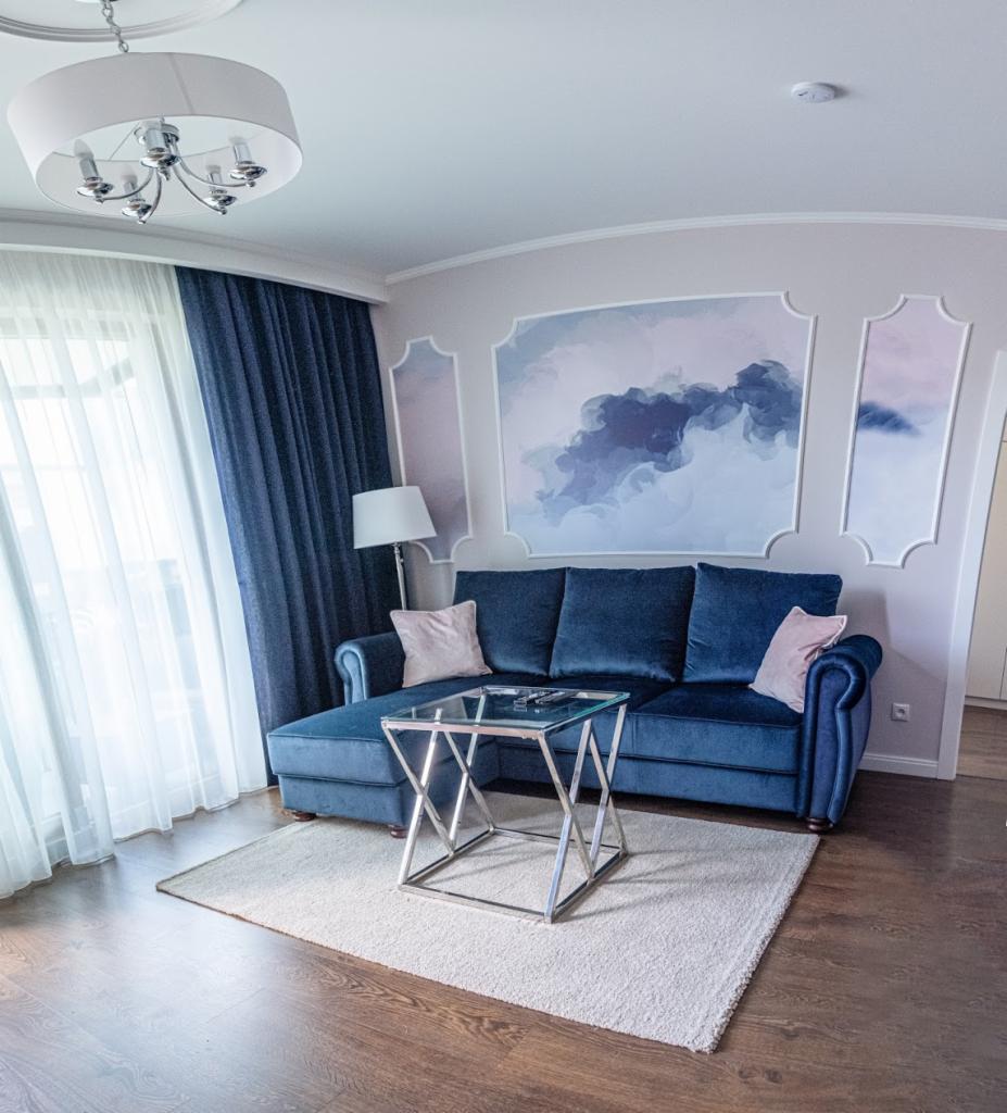 Salon z granatową sofą narożną, szklanym stolikiem kawowym na dywanie i dekoracyjną ścianą z motywem chmur w ramkach.