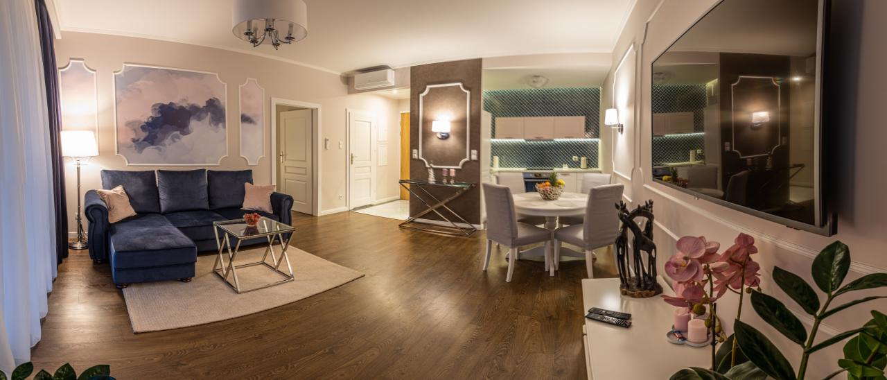 Panoramiczne ujęcie salonu z aneksem kuchennym: granatowa sofa z poduszkami, szklany stolik kawowy na beżowym dywanie, okrągły stół z krzesłami w jadalni, dekoracje ścienne w formie obrazów...