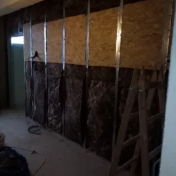 Montaż izolacji z wełny mineralnej i płyt OSB na ścianie z metalowym stelażem, widoczne kable elektryczne i drabina drewniana.