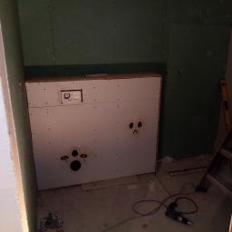 Wykonczymy Was - Stelaż podtynkowy do WC zabudowany płytami gipsowo-kartonowymi na zielonych ścianach, widoczne otwory na instalacje, poziomica i wiertarka na podłodze.