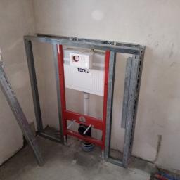 Wykonczymy Was - Stelaż podtynkowy TECE zamontowany w narożniku łazienki, widoczne profile metalowe i przyłącza kanalizacyjne, surowe ściany w tle.