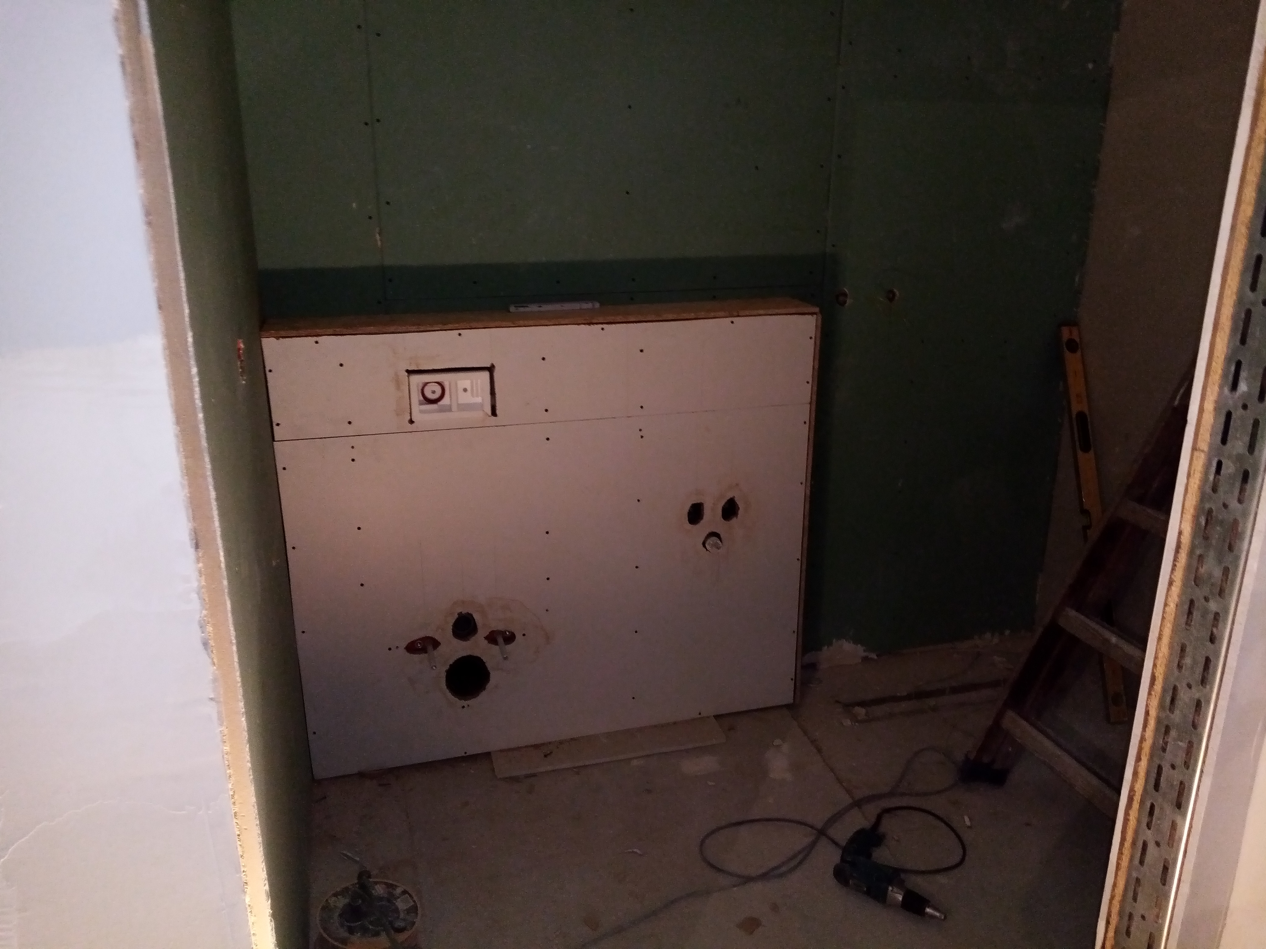 Stelaż podtynkowy do WC zabudowany płytami gipsowo-kartonowymi na zielonych ścianach, widoczne otwory na instalacje, poziomica i wiertarka na podłodze.