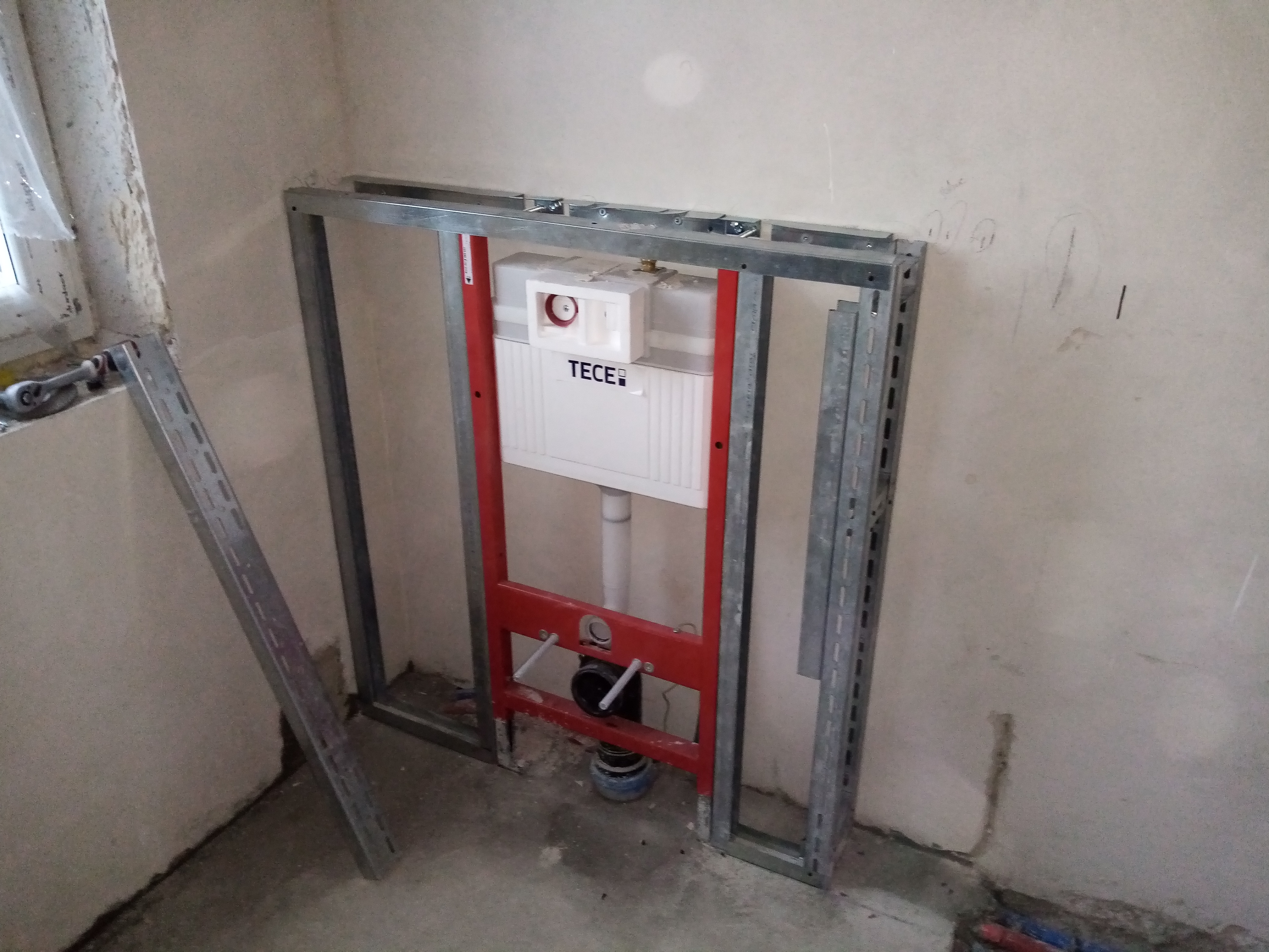 Stelaż podtynkowy TECE zamontowany w narożniku łazienki, widoczne profile metalowe i przyłącza kanalizacyjne, surowe ściany w tle.