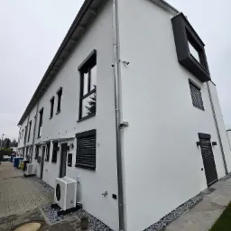 Nowoczesny szeregowy budynek mieszkalny z białymi ścianami, czarnymi ramami okiennymi i roletami zewnętrznymi, widok z poziomu chodnika wyłożonego kostką.