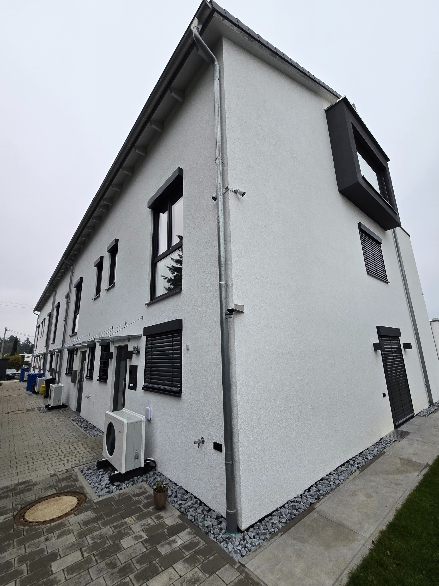 Nowoczesny szeregowy budynek mieszkalny z białymi ścianami, czarnymi ramami okiennymi i roletami zewnętrznymi, widok z poziomu chodnika wyłożonego kostką.