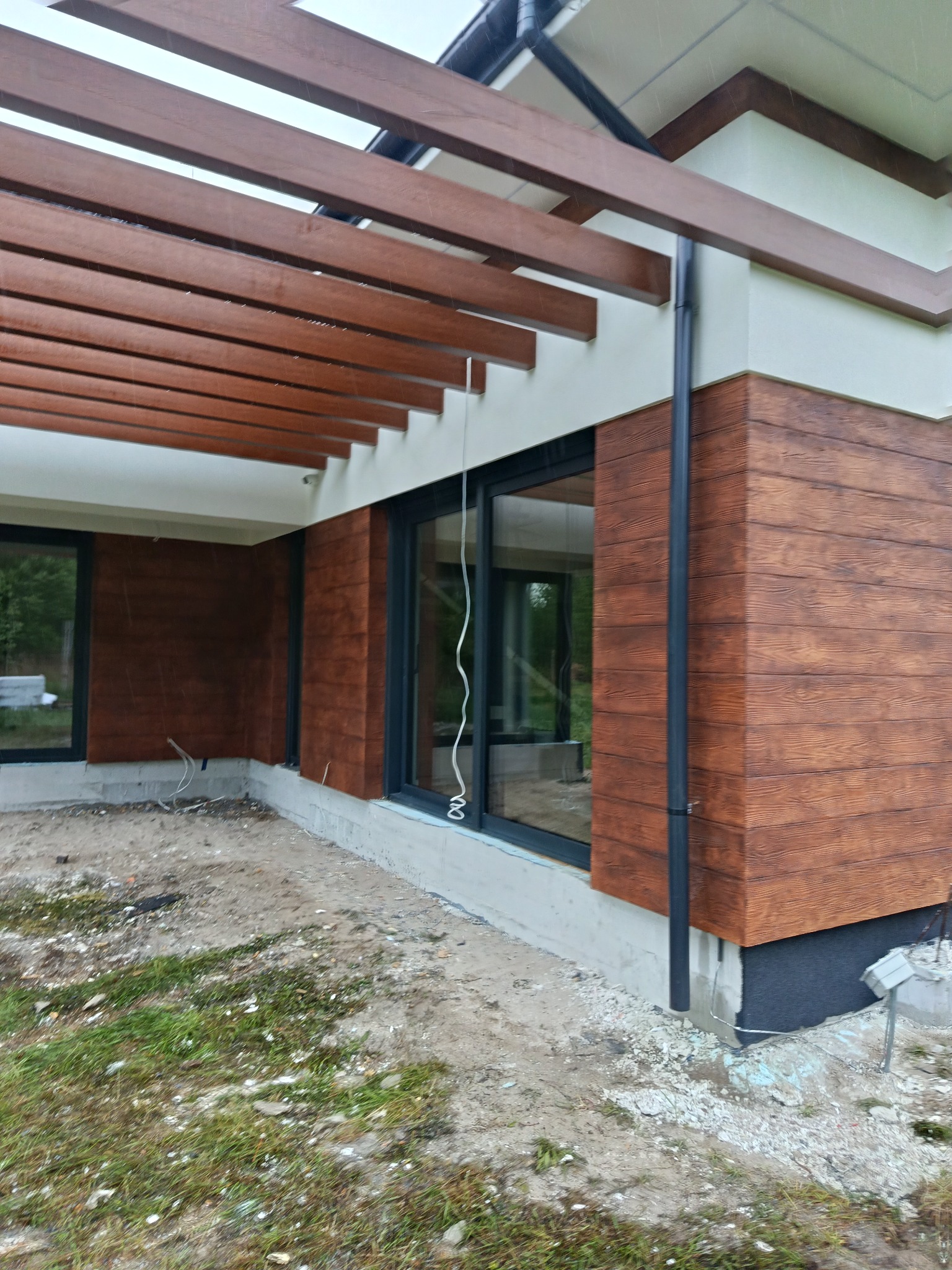 Elewacja domu z brązową okładziną imitującą drewno, czarne rynny i okna, drewniana pergola. Widoczne niedokończone prace ziemne wokół budynku.