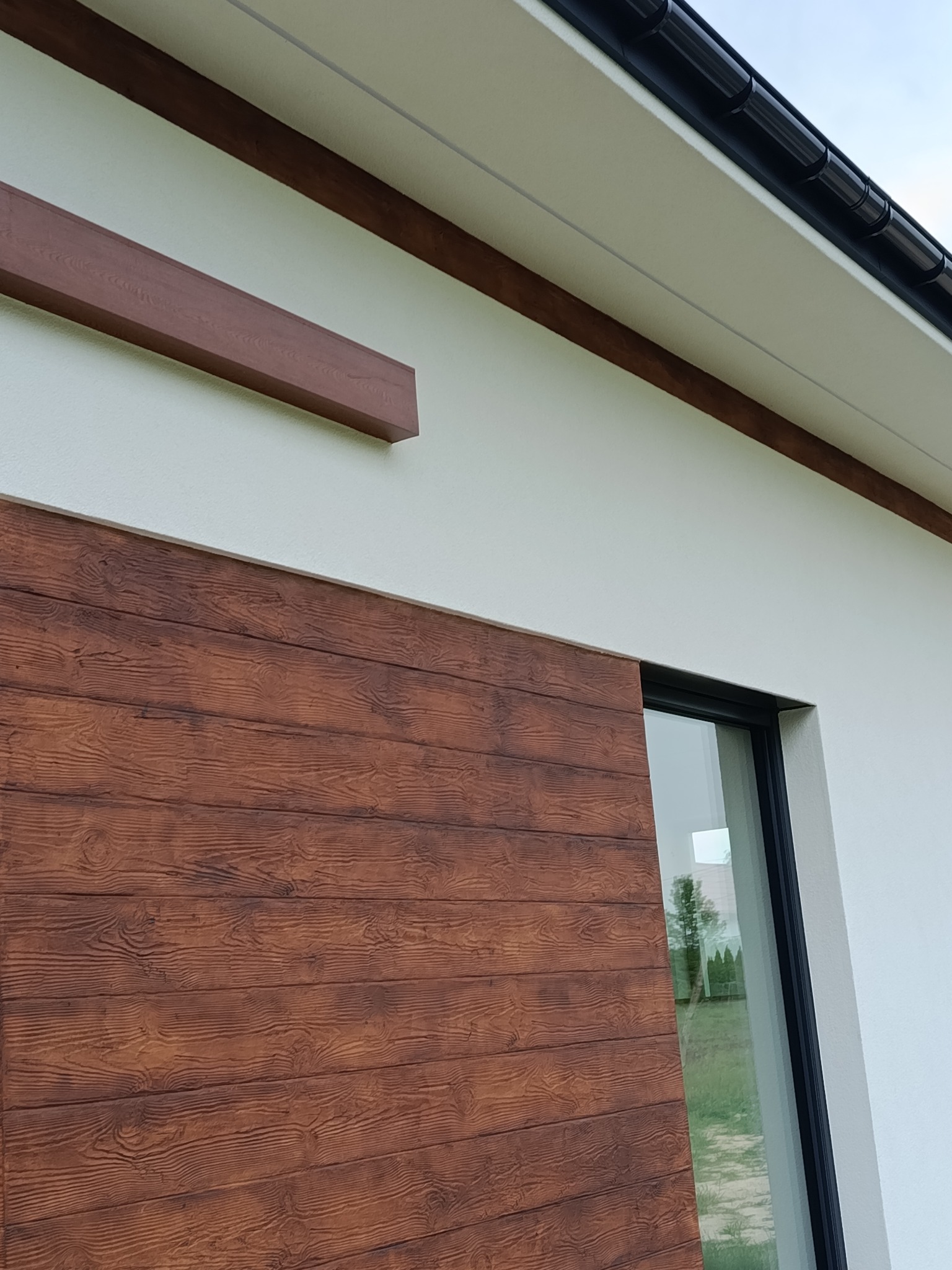 Fragment elewacji domu z drewnopodobną okładziną i oknem, widok z dołu. Elementy dekoracyjne w kolorze brązowym. Nowoczesne wykończenie.