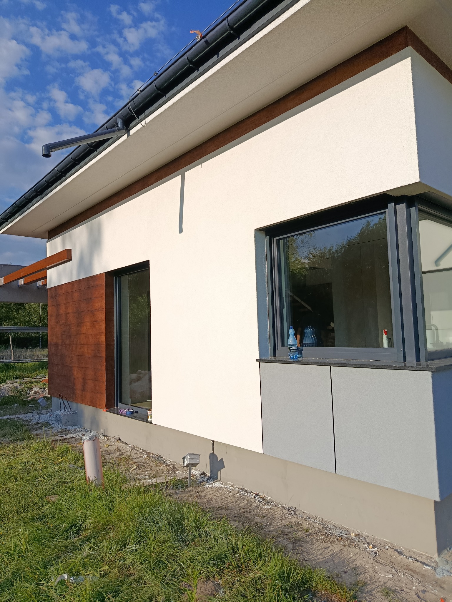 Nowoczesna elewacja domu jednorodzinnego z drewnianą okładziną i szarymi panelami. Widoczne okna z ciemnymi ramami i fragment ogrodu.