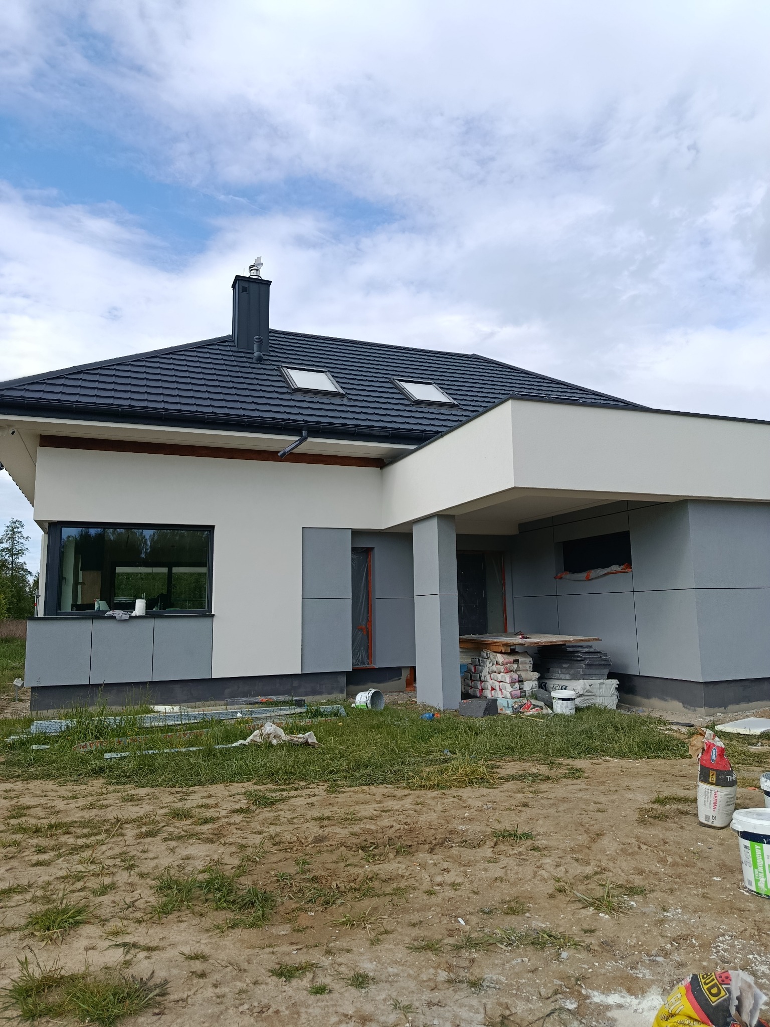 Nowoczesny dom jednorodzinny z szarą elewacją i czarnym dachem, częściowo ocieplony, z widocznymi materiałami budowlanymi na trawniku.