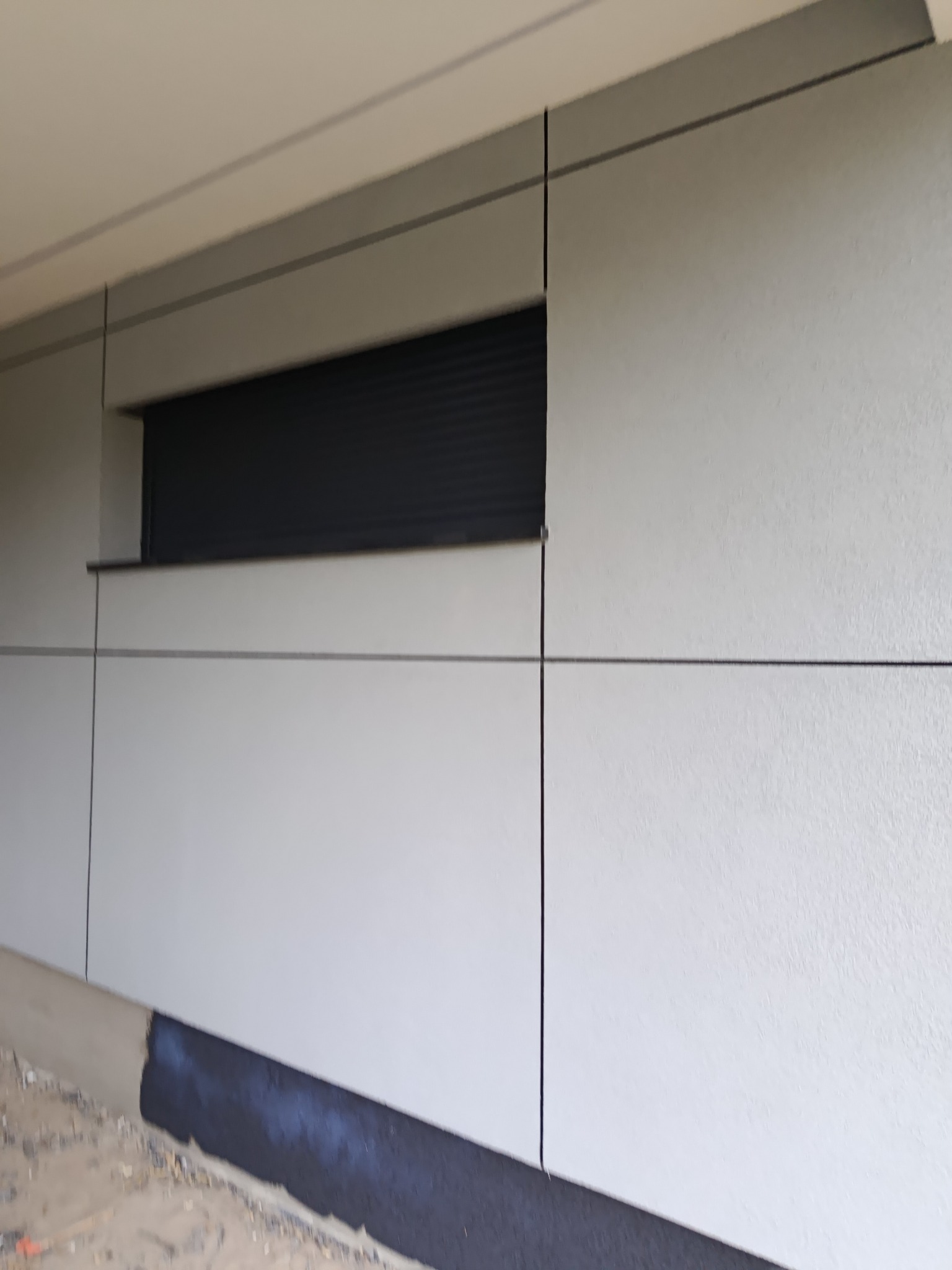 Elewacja budynku wykończona panelami w odcieniach szarości, z poziomymi i pionowymi fugami. Widoczne okno z żaluzjami i fragment cokołu.