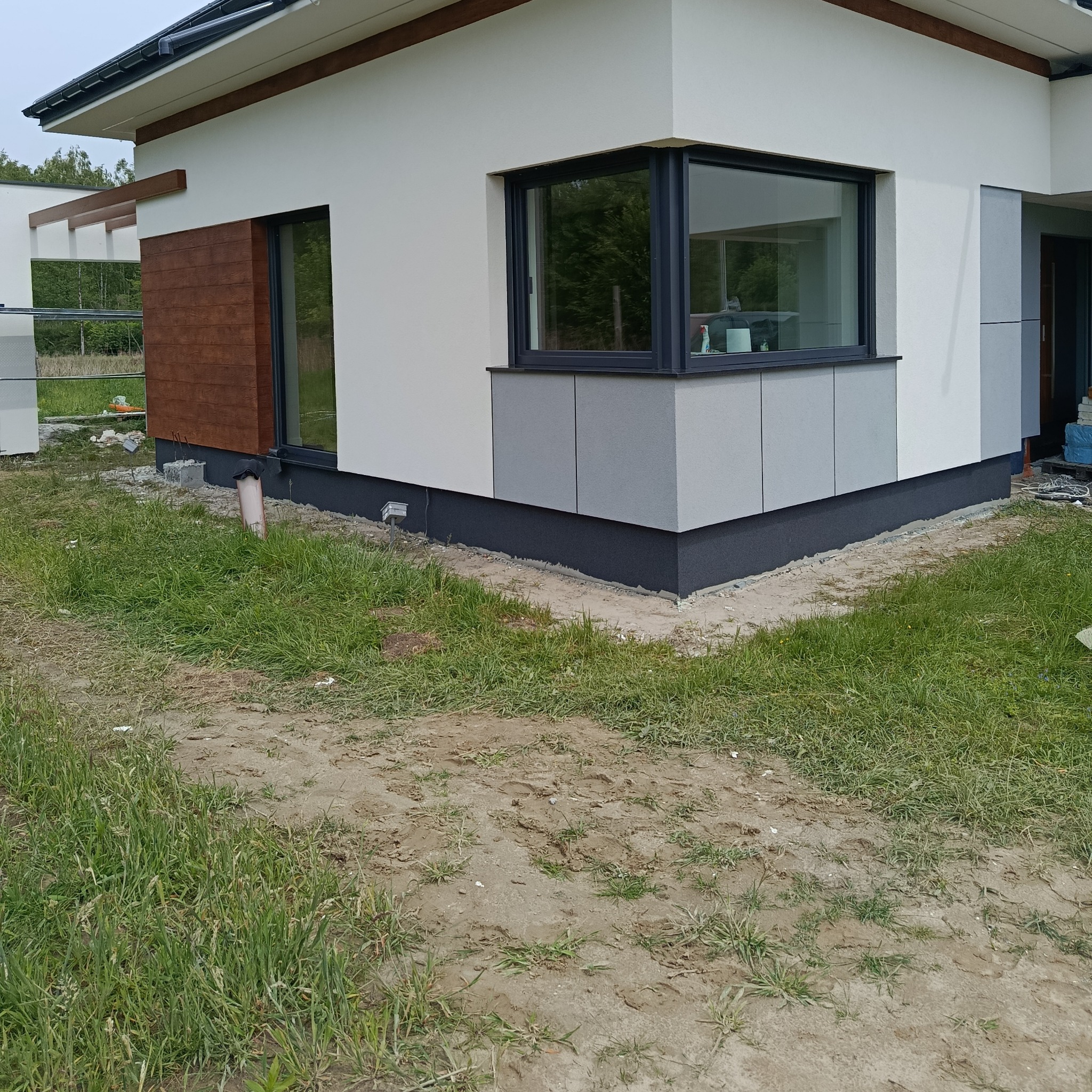 Fragment elewacji domu z drewnianą okładziną i szarymi panelami. Widoczne okno narożne i trawnik. Detal wykończenia budynku.