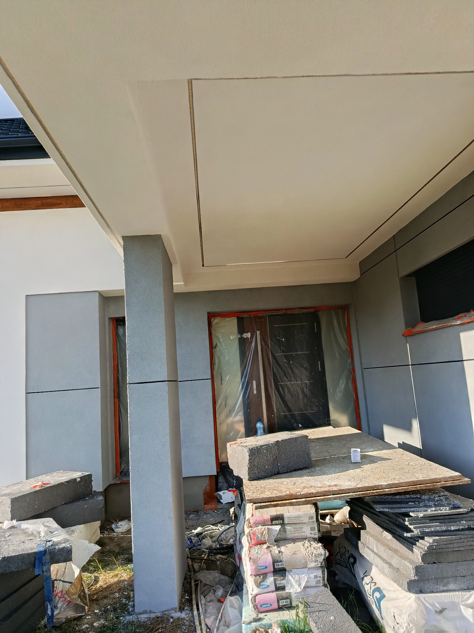 Wykończony fragment elewacji domu z szarą fasadą i białym stropem, widoczne materiały budowlane i narzędzia.