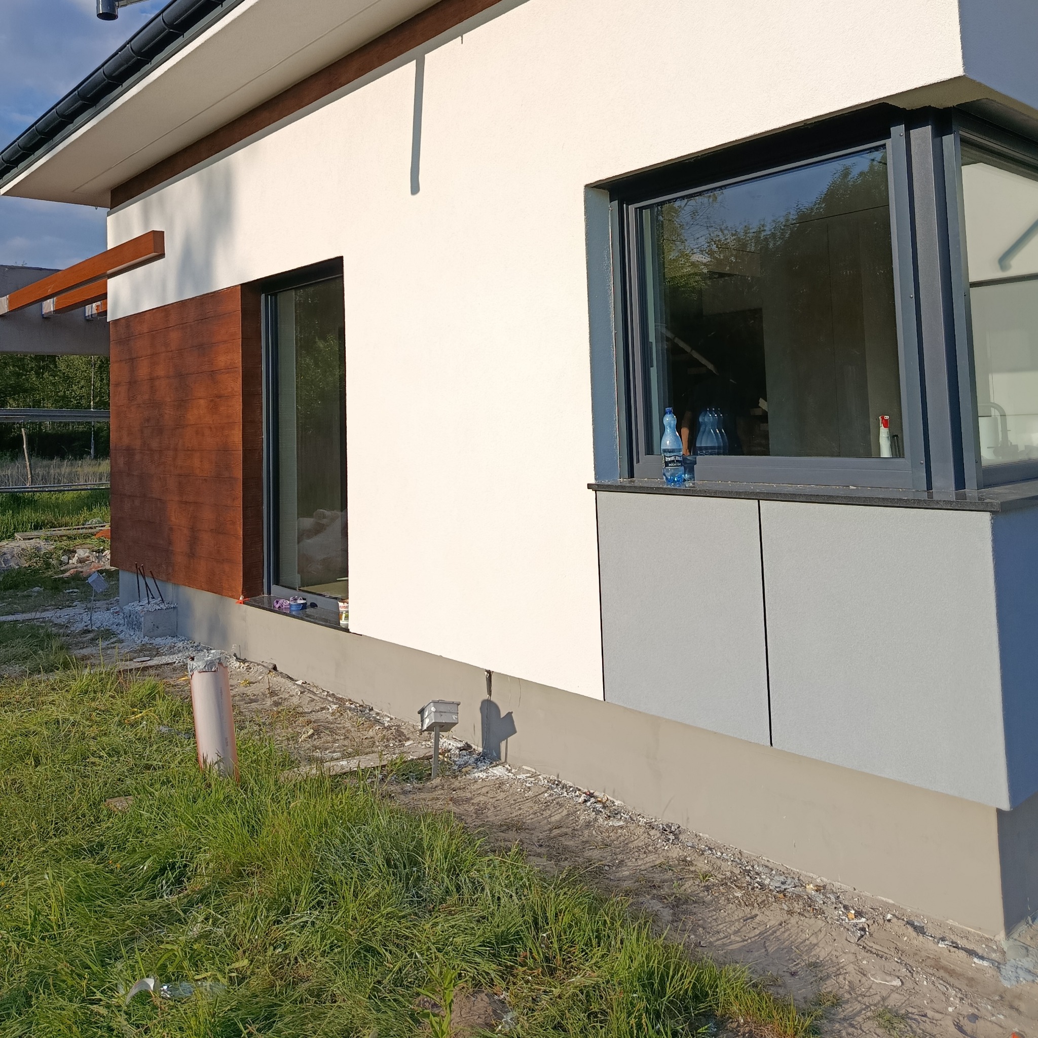 Fragment elewacji domu z oknami, wykończony drewnopodobną okładziną i szarą płytą. Widoczne trawnik i elementy konstrukcyjne.