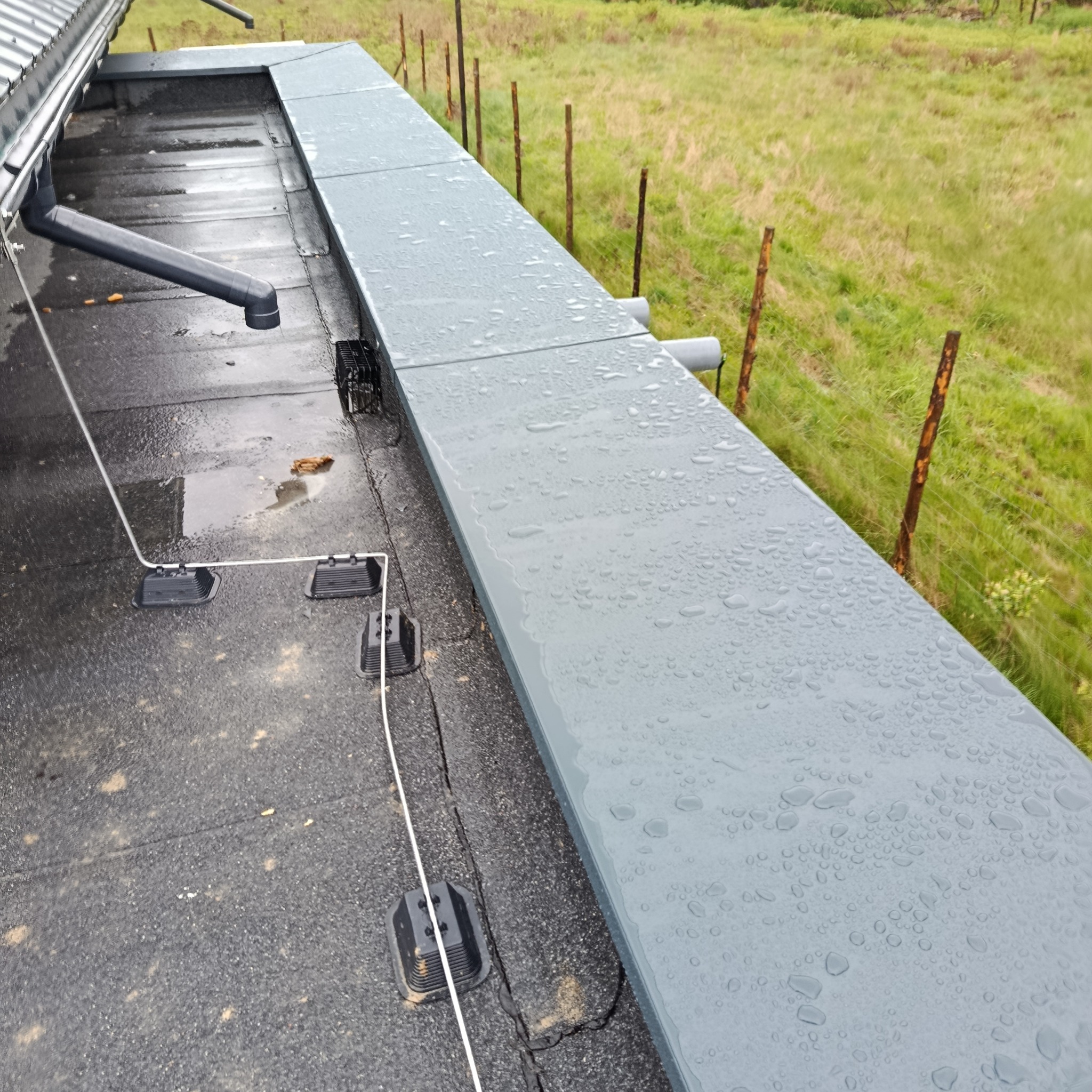 Widok z góry na fragment dachu pokrytego papą z systemem odprowadzania wody. Widoczne elementy mocujące, rury spustowe i obróbki blacharskie. Na powierzchni widoczne krople deszczu.