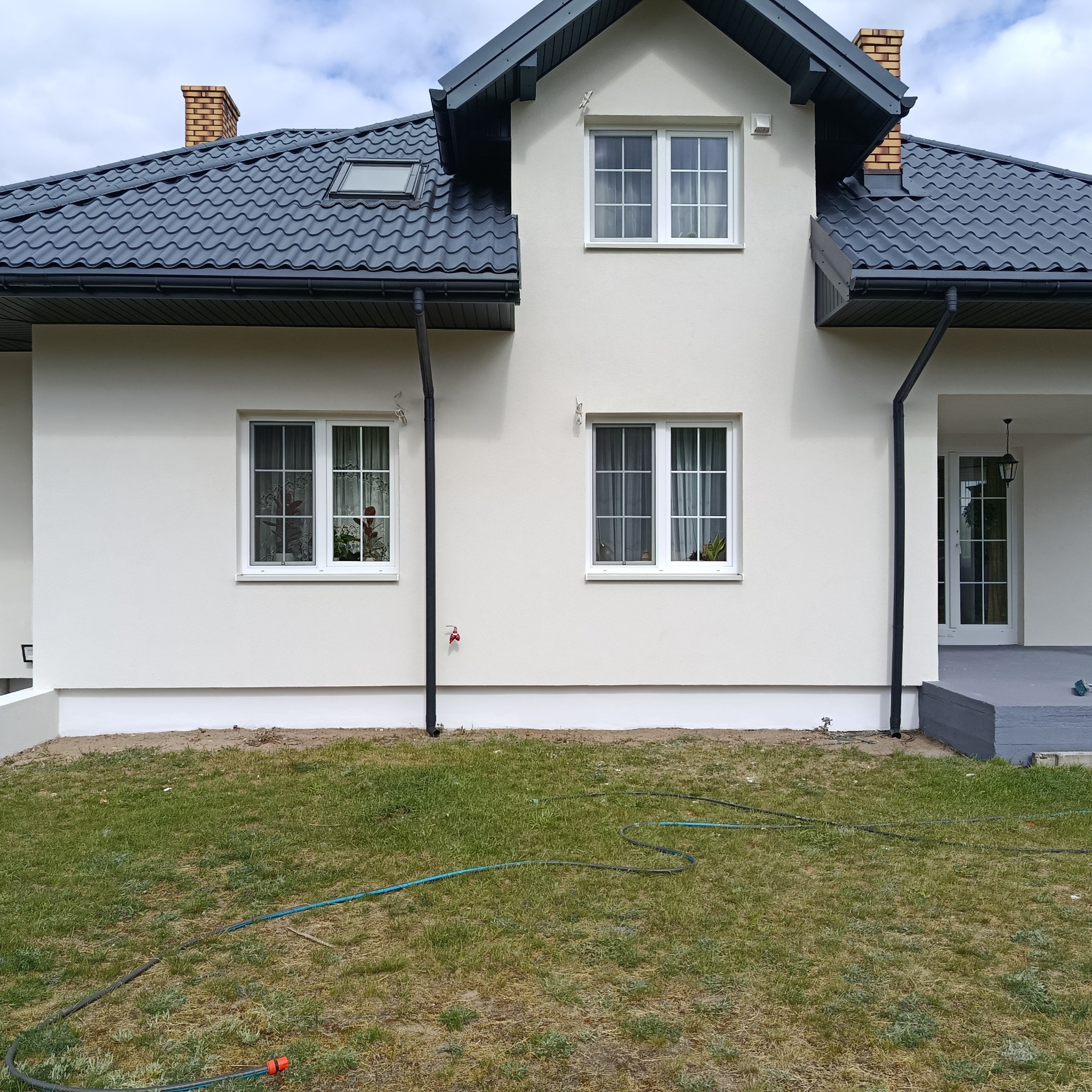 Otynkowany dom jednorodzinny z ciemną dachówką i białymi oknami. Widoczne rynny, fragment trawnika z wężem ogrodowym. Świeżo pomalowana elewacja.