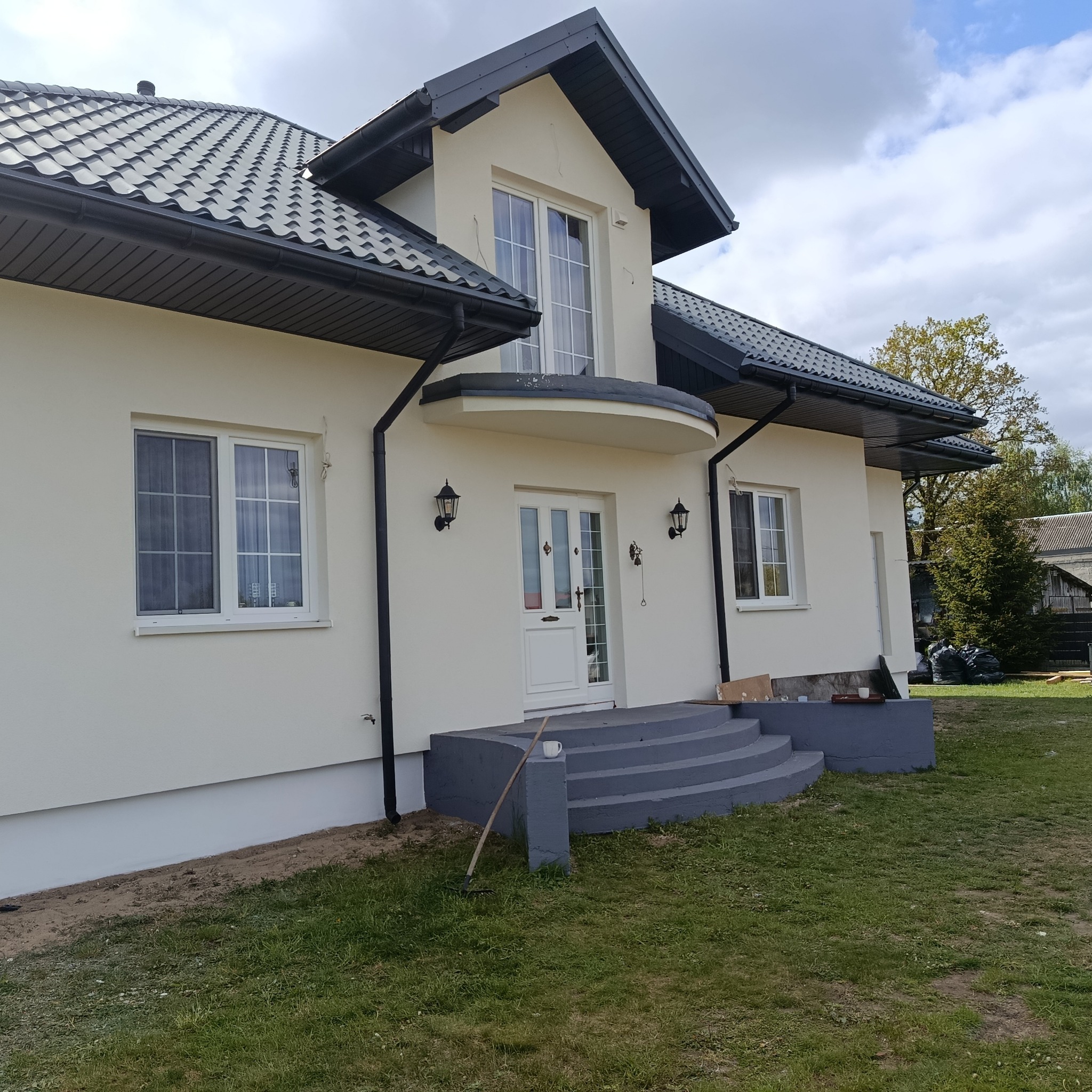 Elewacja domu jednorodzinnego po remoncie. Jasna fasada, ciemna dachówka, okna z białymi ramami. Widoczne schody wejściowe i fragment trawnika. Słoneczny dzień.