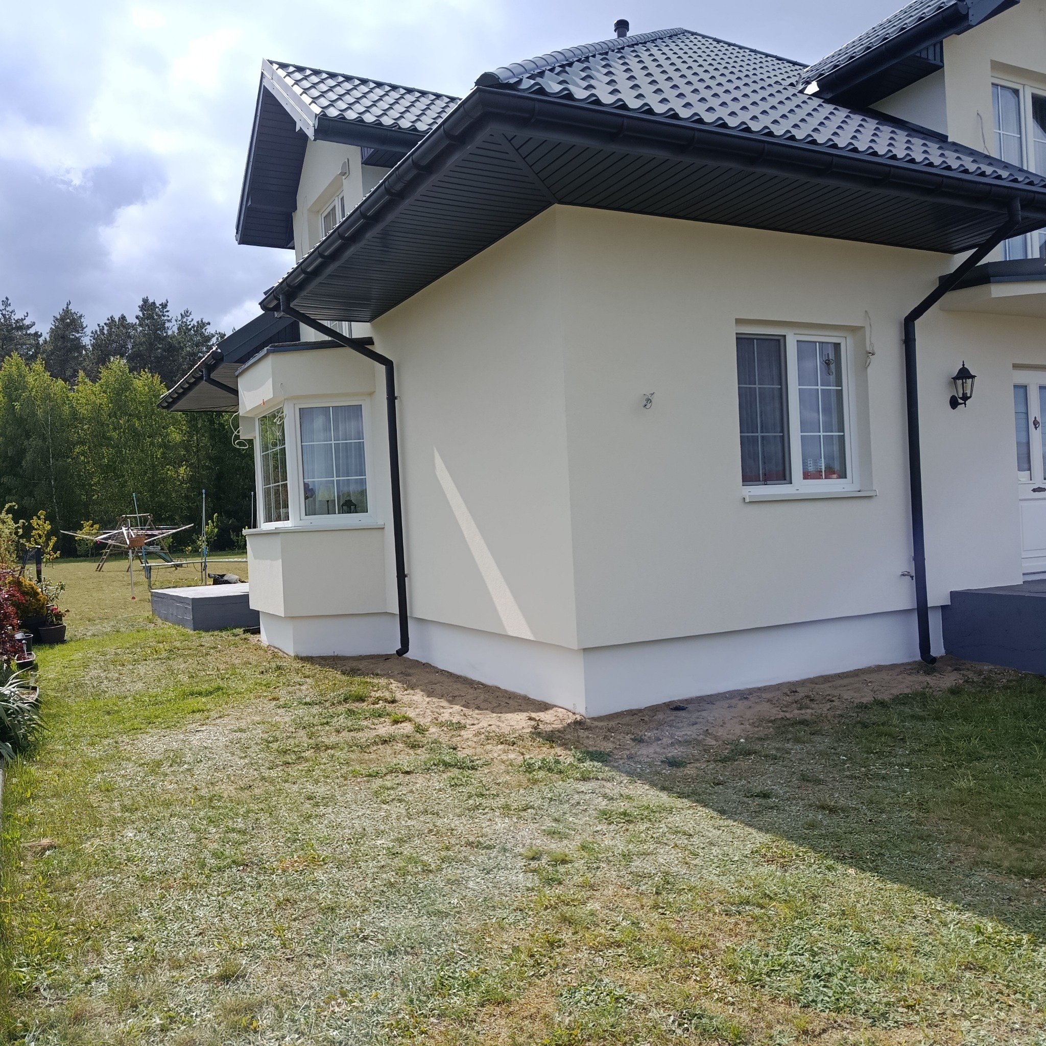 Elewacja domu jednorodzinnego z oknami i czarną dachówką, po remoncie. Widok z zewnątrz, trawnik w pierwszym planie.