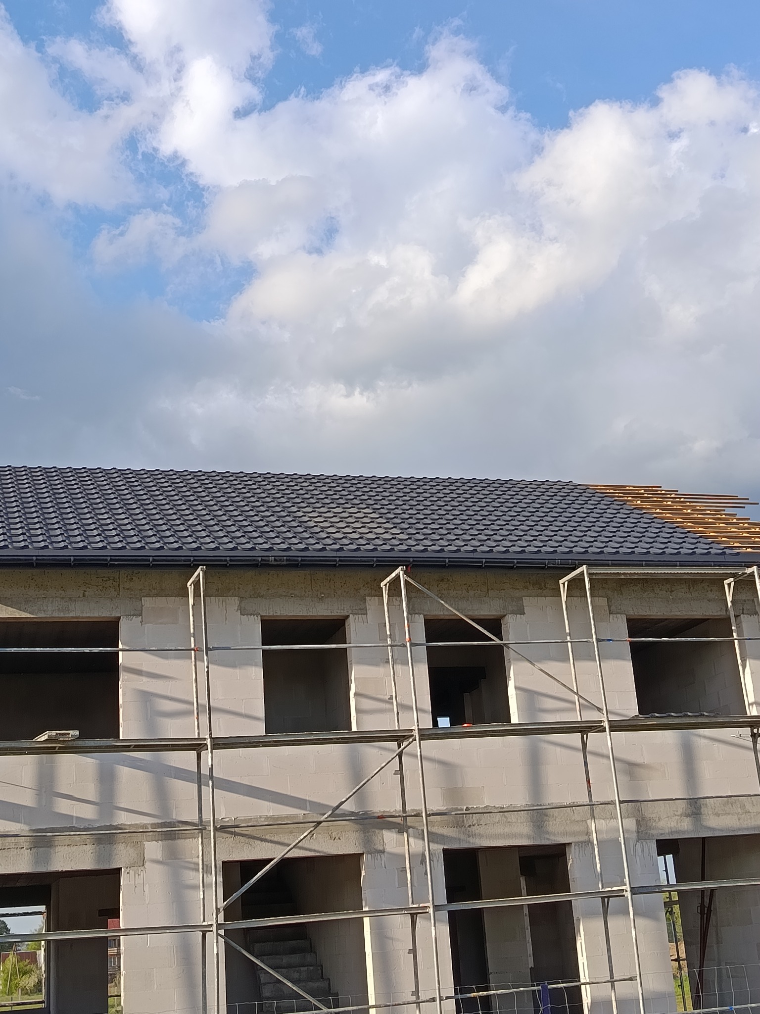 Budowa domu z białych bloczków, widoczne rusztowania i fragment dachu z czarnej dachówki na tle nieba z chmurami. Częściowo położone łaty pod dach.