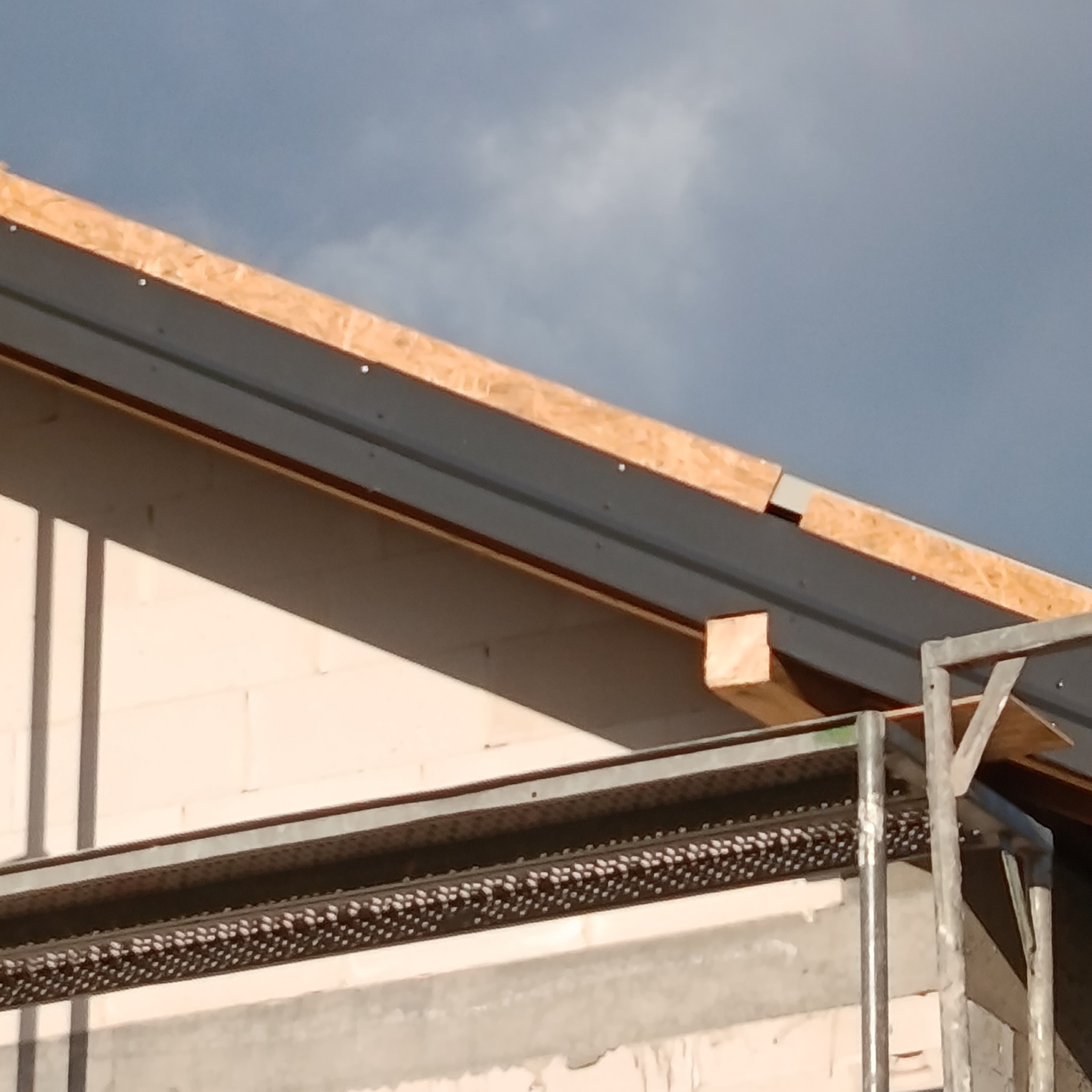 Fragment elewacji budynku w trakcie ocieplania, widoczne rusztowanie i elementy konstrukcyjne dachu. Jasne bloczki i drewniane elementy konstrukcji.