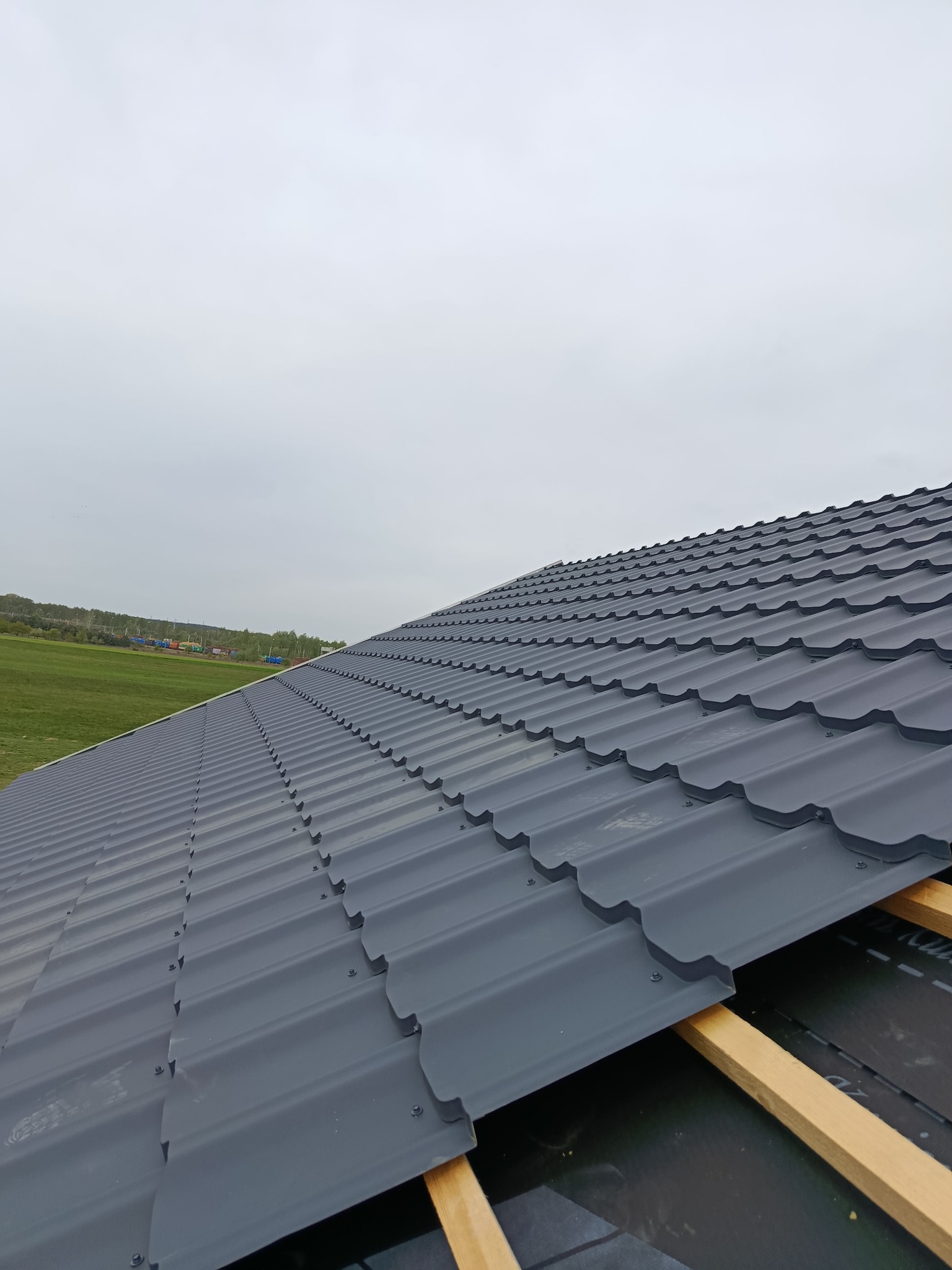 Szary dach z blachy trapezowej, widok z góry, częściowo zamontowany na drewnianej konstrukcji. W tle zielone pole i pochmurne niebo.