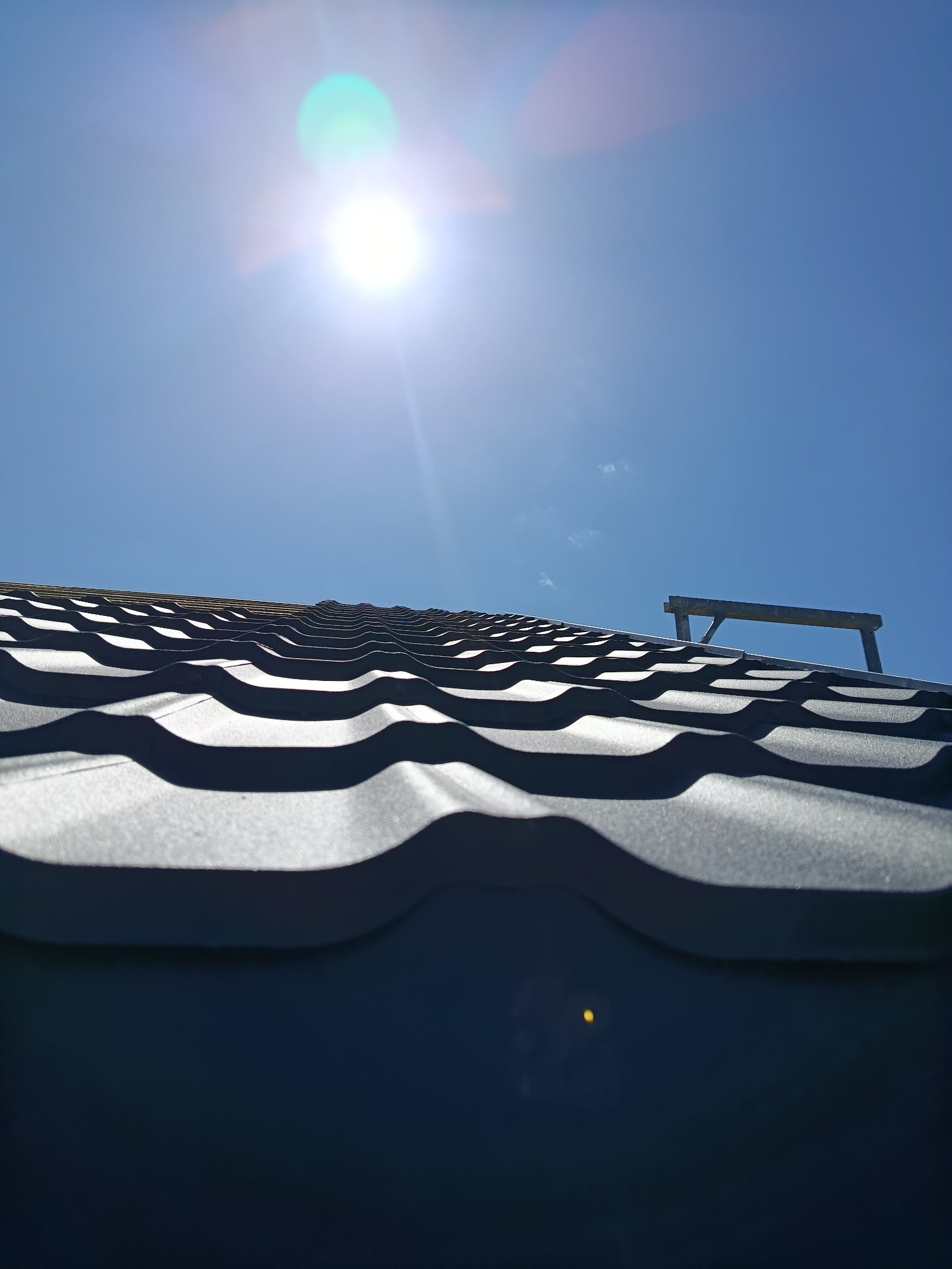 Błękitne niebo nad ciemną, metalową dachówką w jodełkę. W tle widoczny fragment rusztowania i oślepiające słońce.
